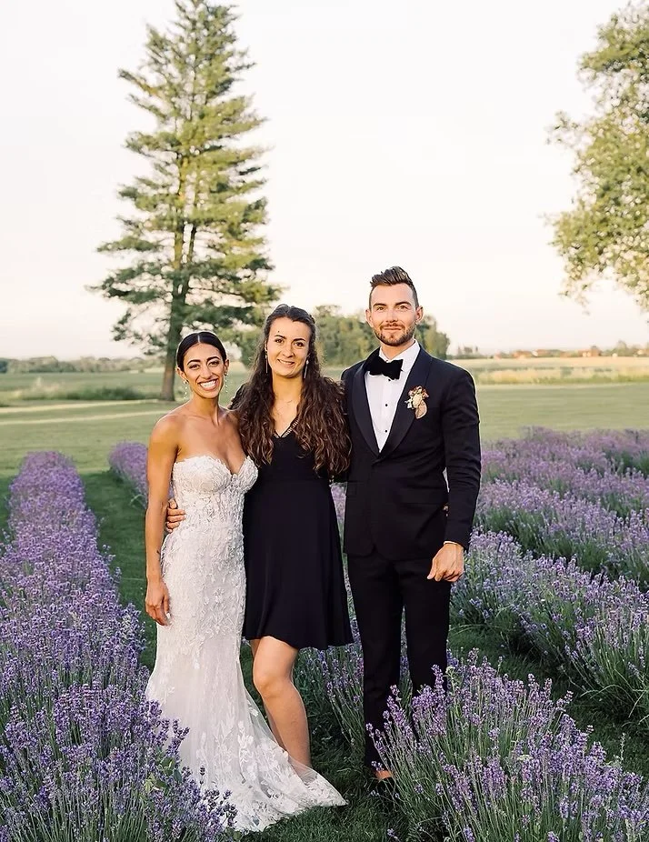 A bride, a woman, and a groom standing outdoors in a lavender field, with trees and open landscape in the background, during a wedding celebration.