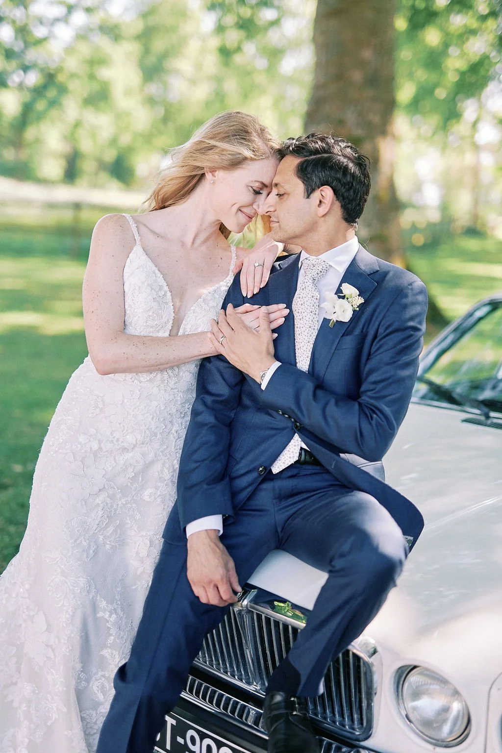 A bride in a white lace wedding dress and a groom in a blue suit with a white shirt and tie, both smiling with foreheads touching and hands clasped, standing outdoors next to a white luxury car in a lush green park.