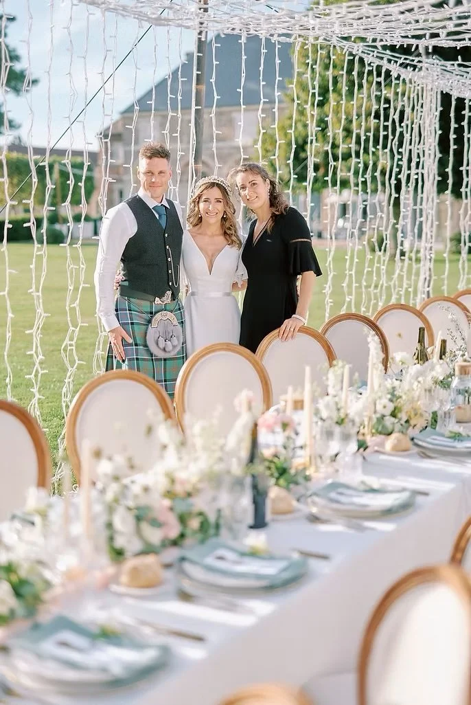Three people standing close together behind a decorated outdoor banquet table at a wedding reception.