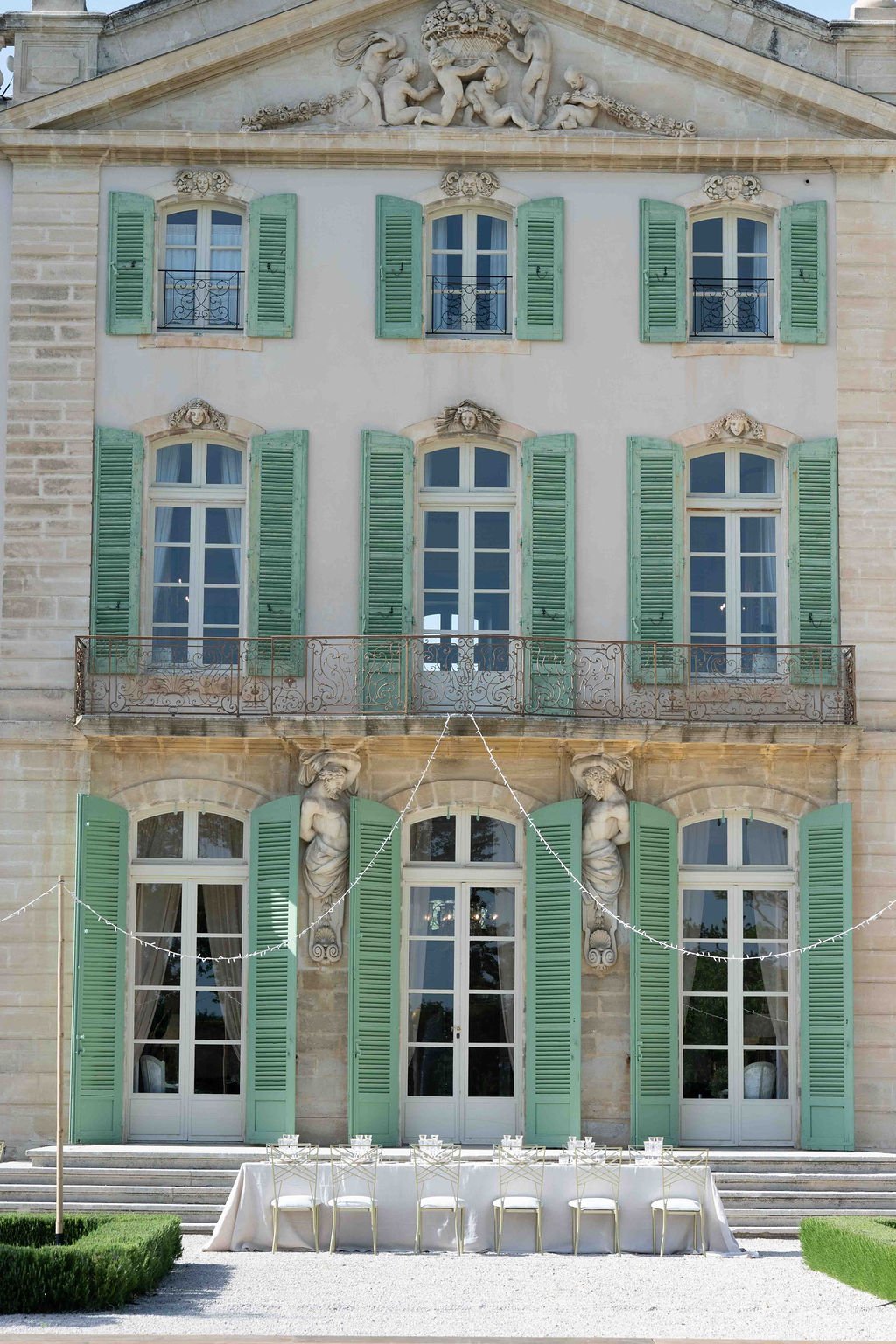 The front of a historic building featuring six large windows with green shutters and ornate stone sculptures. There are three windows on each of the two upper floors, and a balcony with decorative iron railings. The ground level has three large glass