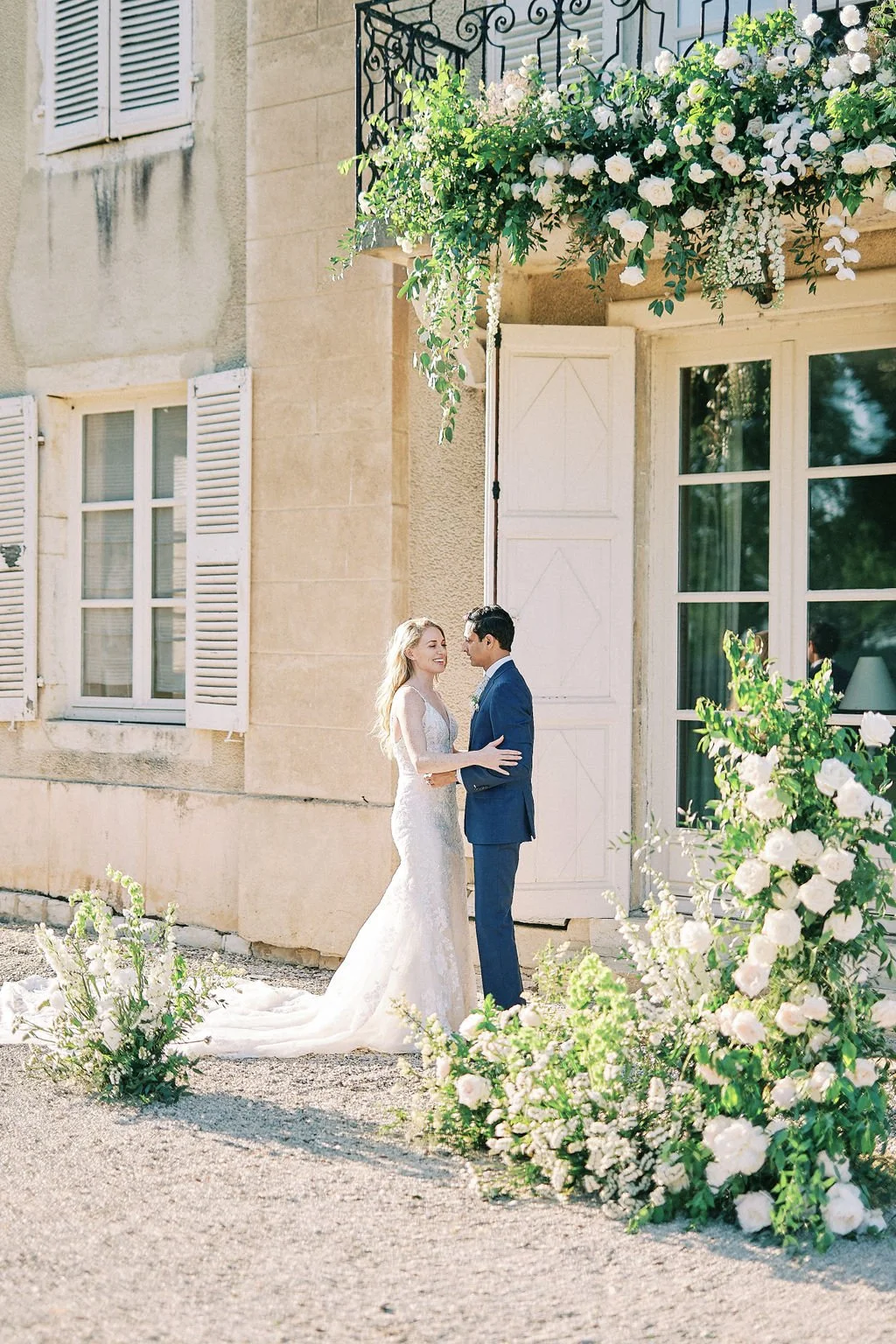 A bride and groom stand close together outside a building with white shutters and large windows, surrounded by flowering plants and greenery.