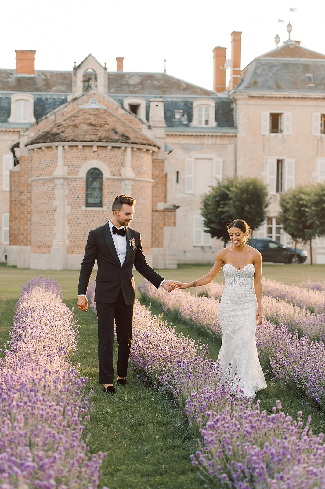 A bride and groom holding hands and walking through lavender rows outdoors in front of a historic building, with the groom in a black tuxedo and the bride in a strapless wedding gown.