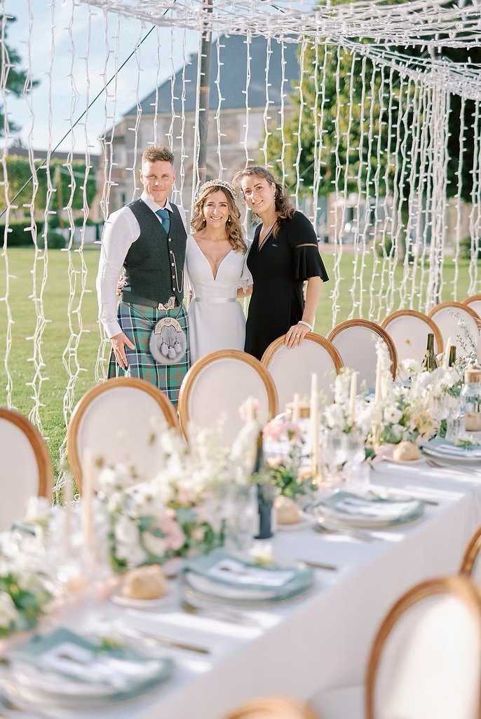 Three people standing behind a decorated wedding reception table outdoors with a string light backdrop, including a bride in a white dress, a groom in traditional Scottish attire, and a woman in a black dress.