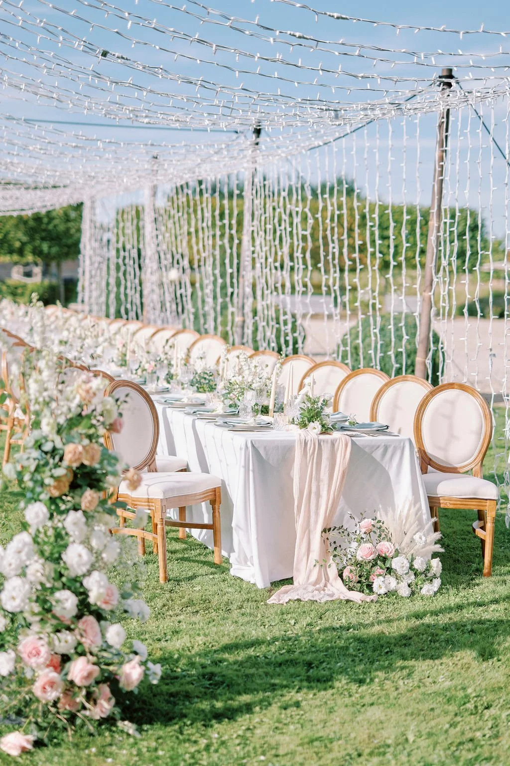 Outdoor wedding reception with a long table decorated with white flowers, greenery, and a blush pink table runner, set under string lights and a decorative net canopy, on a grassy area with a soft natural background.
