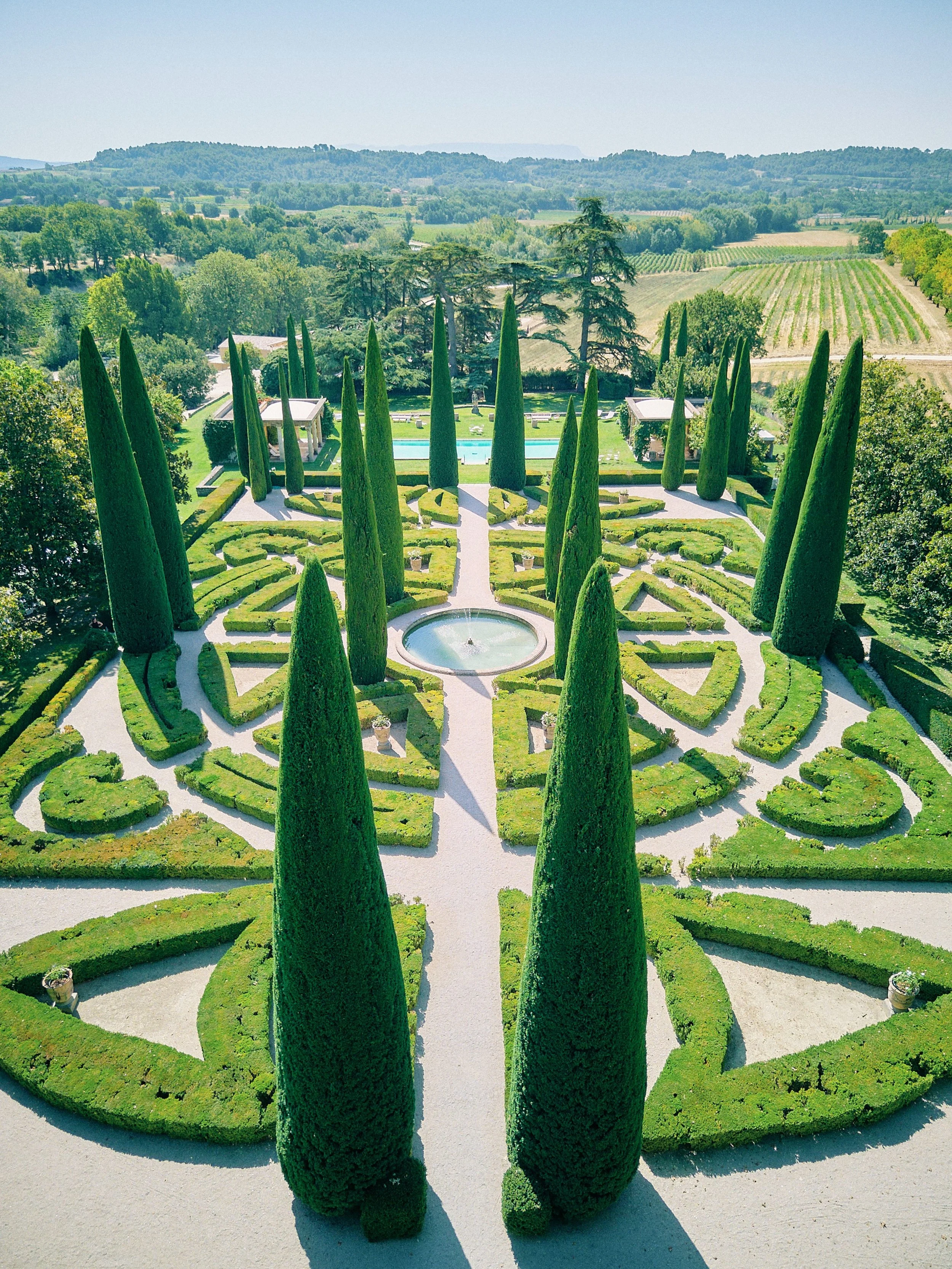 Aerial view of a well-maintained formal garden with tall cypress trees, symmetrical hedges, a central fountain, and surrounding pathways, set against a backdrop of rolling countryside and trees.