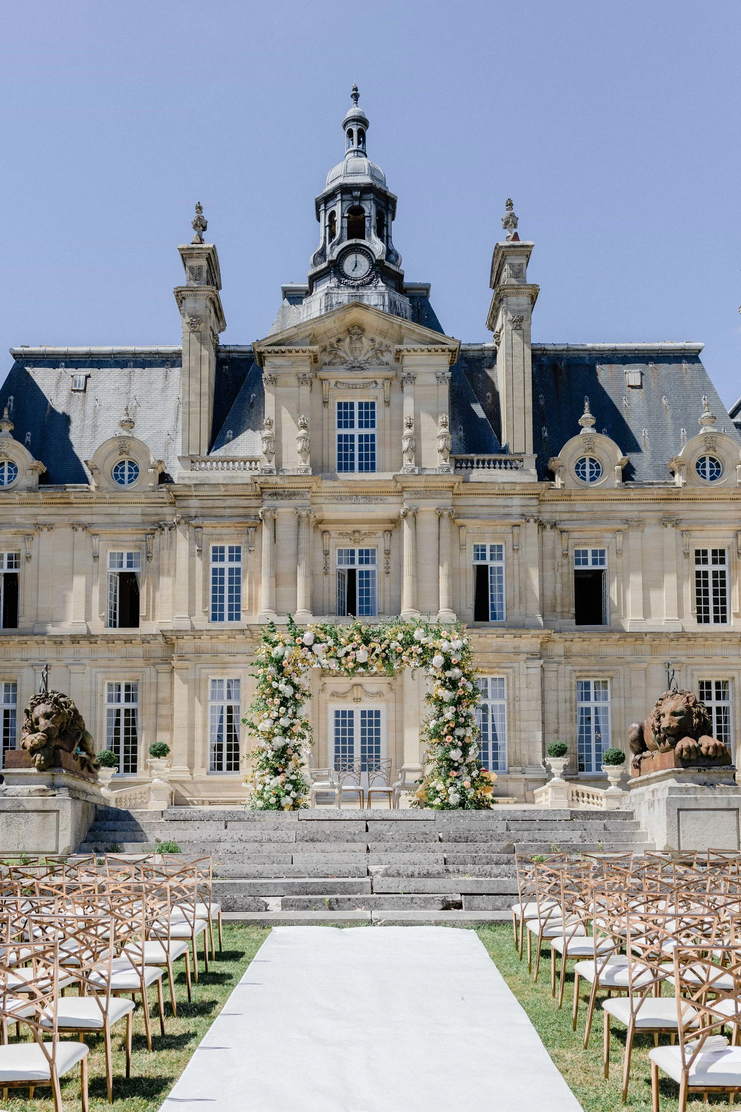 A wedding ceremony setup in front of a grand, historic building with a clock tower, featuring chairs arranged along a white aisle, and a floral arch at the top of the stairs.