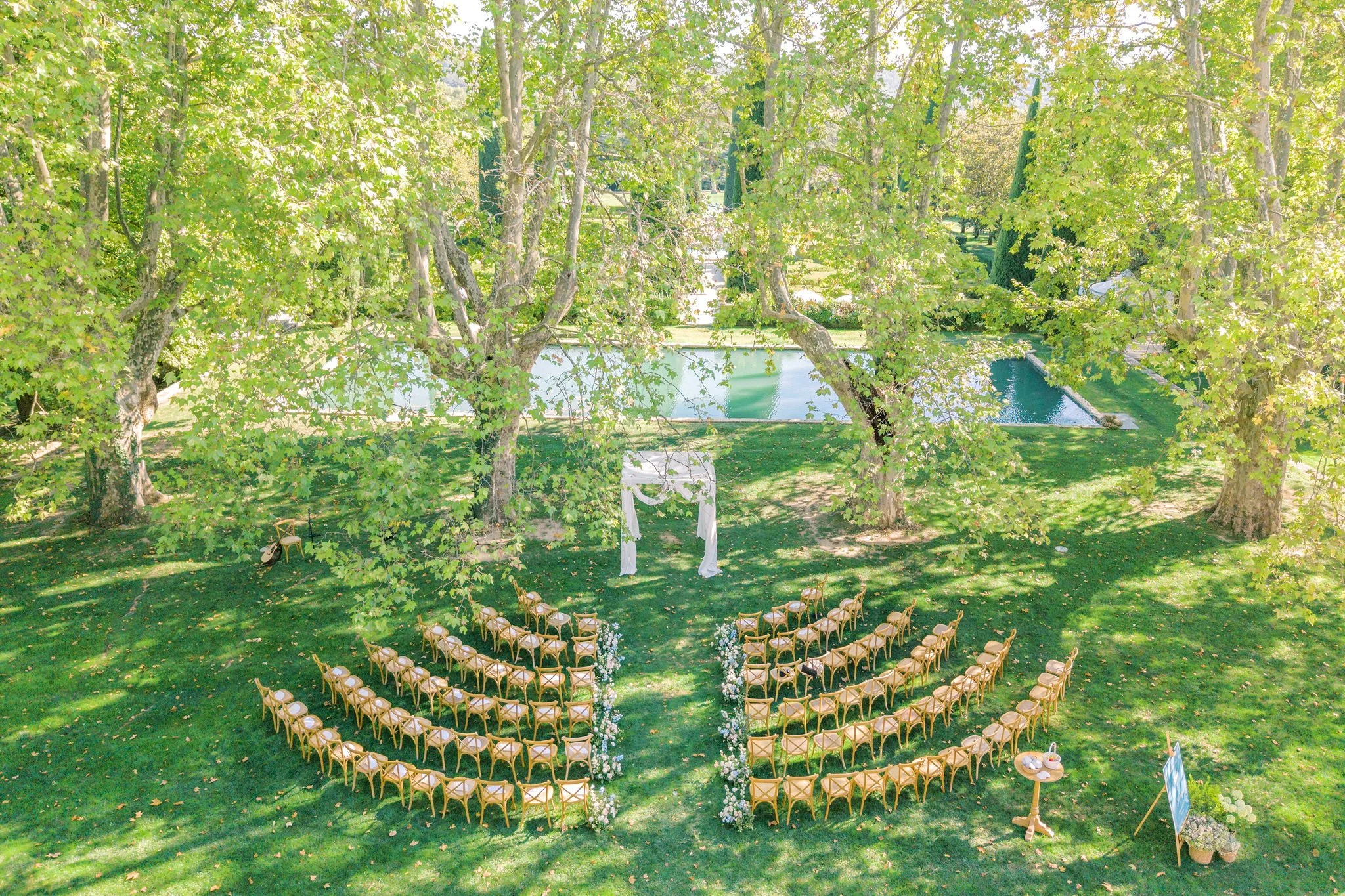 Outdoor wedding setup with rows of chairs arranged in front of a white floral arch, surrounded by trees and a pond in the background.