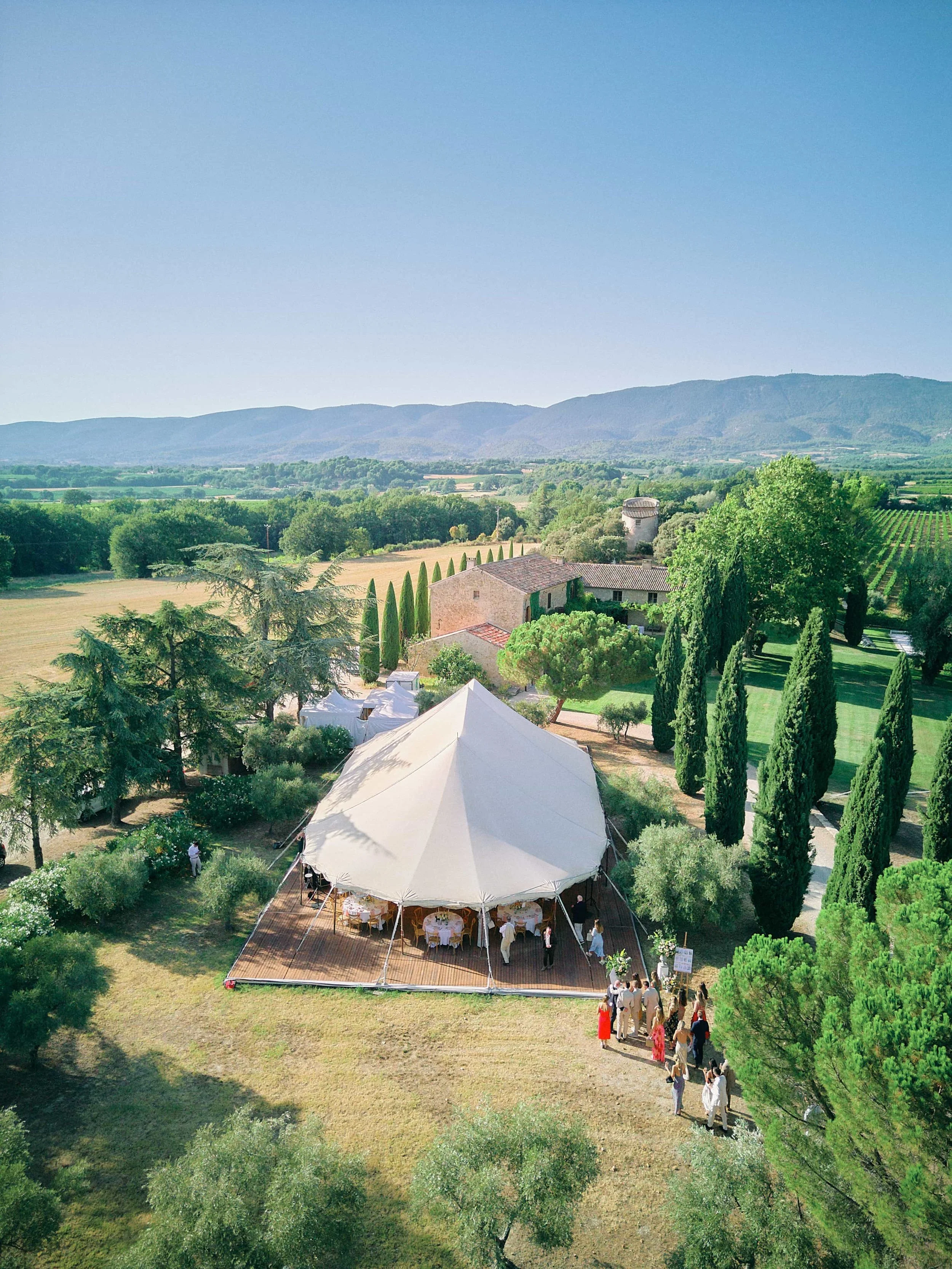 Aerial view of an outdoor event with a large tent set up on grass, surrounded by lush trees and greenery, with people gathered near the entrance. In the background, there are fields, hills, and mountains under a clear blue sky.