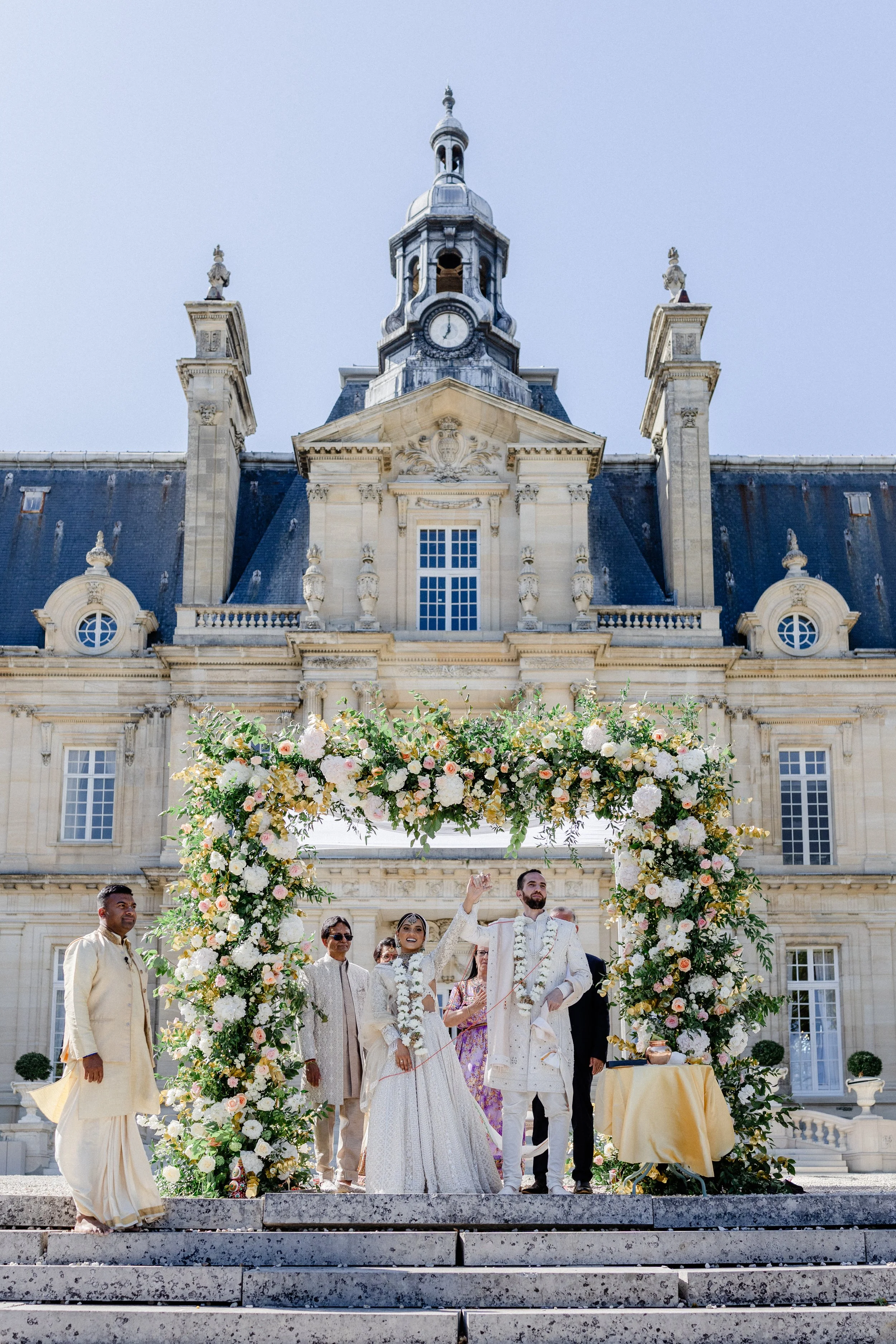 A wedding ceremony taking place outdoors in front of a historic building with a clock tower, under a floral archway with the bride and groom surrounded by guests.