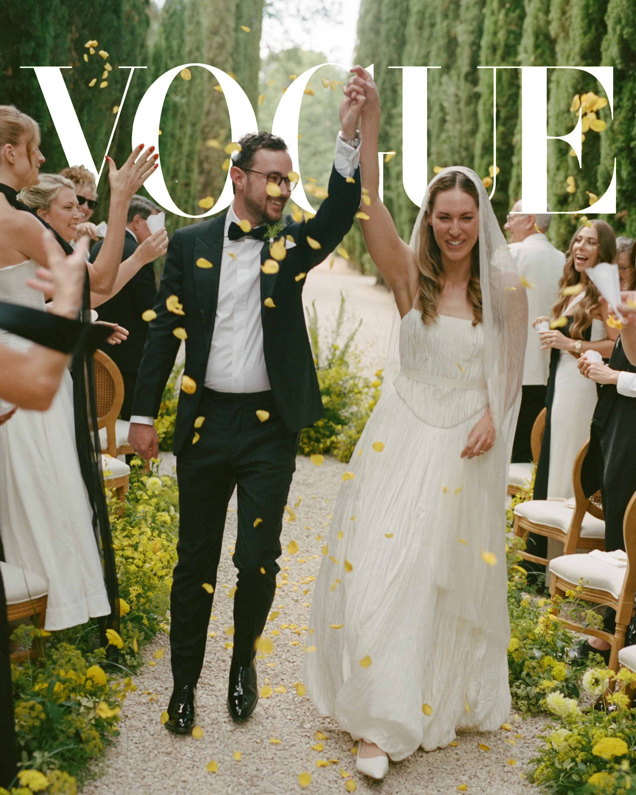 A newlywed couple walking down the aisle, smiling, with guests on both sides throwing yellow flower petals.