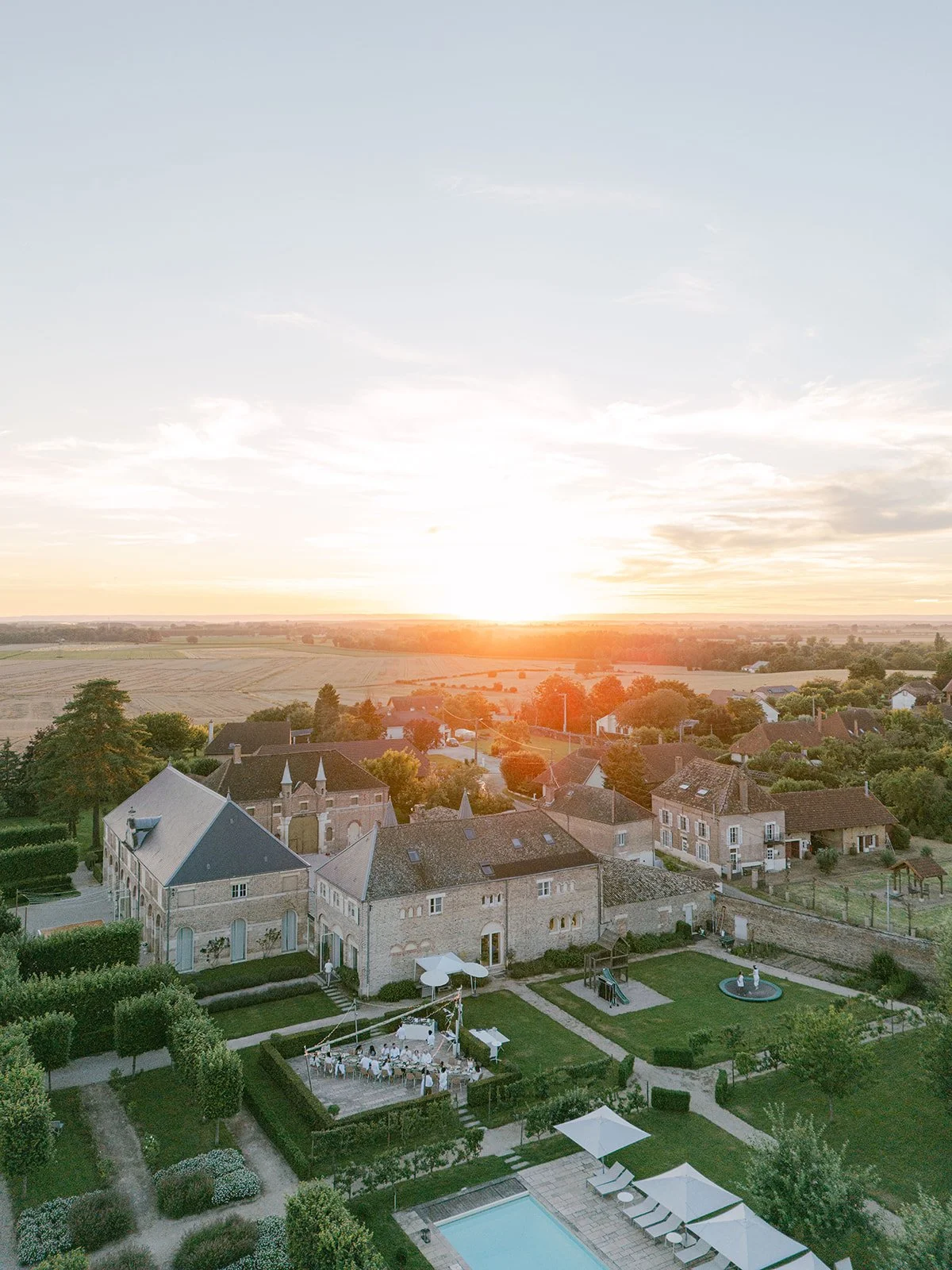 An aerial view of a large house with a landscaped yard, a swimming pool with lounge chairs, and a wedding setup with tables and umbrellas at sunset.