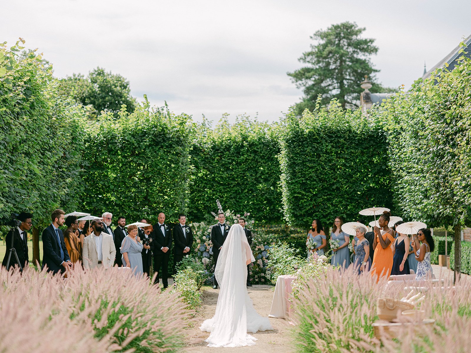 A wedding ceremony outdoors with a bride in a white dress and veil standing in front of guests and bridesmaids in blue and orange dresses holding umbrellas, surrounded by tall green hedges and pink foliage.