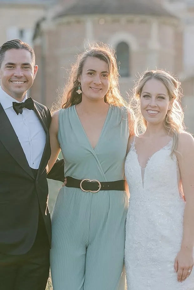 Three people dressed in formal attire standing outdoors in front of a building, smiling at the camera.