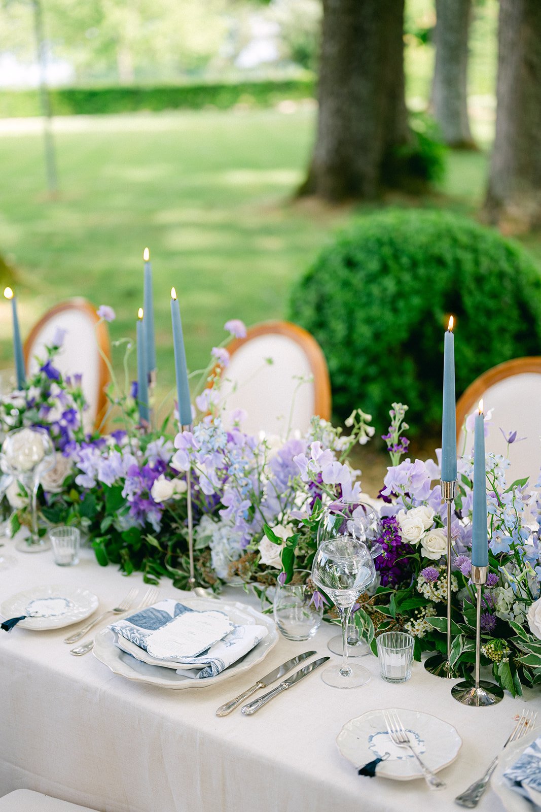 Elegant outdoor table setting with a floral centerpiece, tall candles, and place settings including plates, napkins, and glassware, with trees and grass in the background.