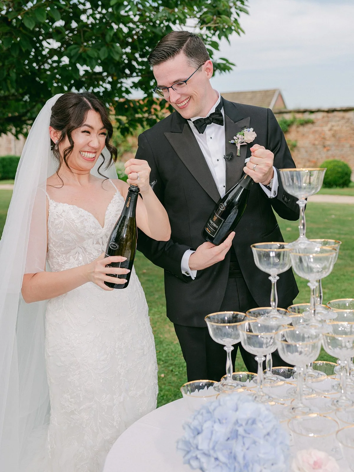 A bride and groom celebrating with champagne at their wedding reception outdoors, smiling and holding champagne bottles, with a pyramid of champagne glasses in front of them.