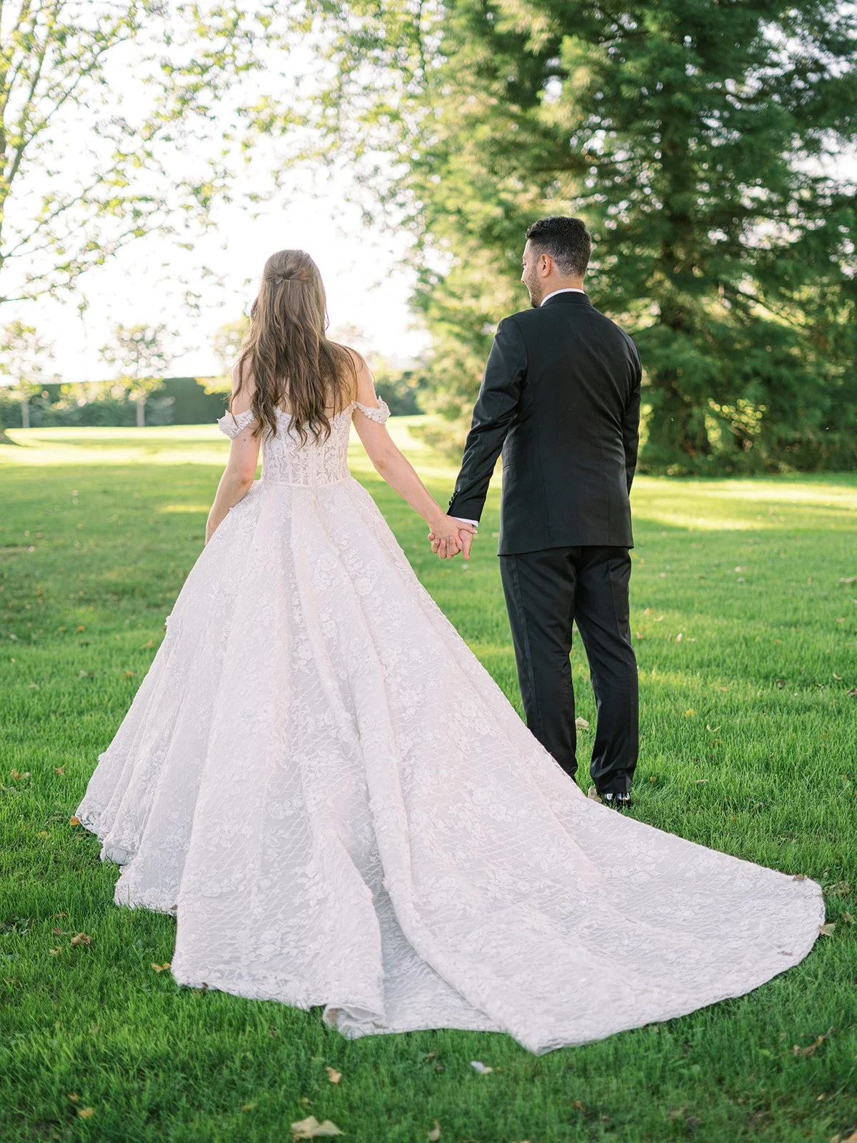 A bride and groom holding hands and walking on a grassy field with trees in the background, during a wedding photoshoot.