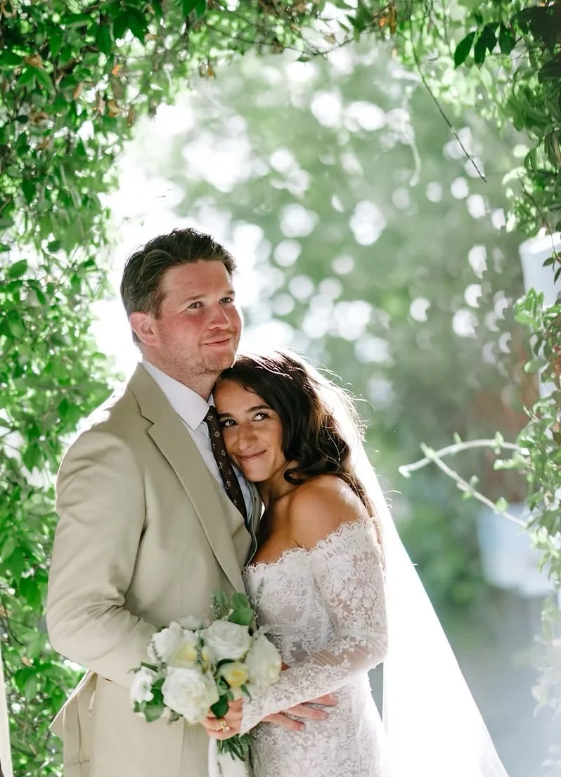 A bride and groom smiling inside a leafy, green arch during their wedding.