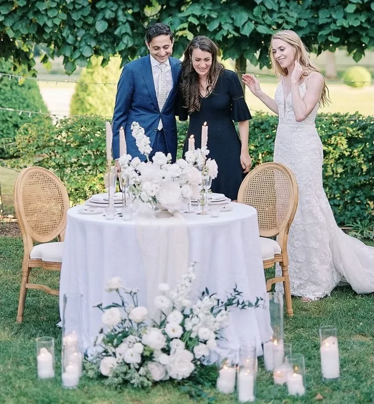 Three people stand around a decorated table outdoors, with a bride in a white wedding gown on the right, a man in a blue suit on the left, and a woman in a dark dress in the middle, all smiling and looking at the table. The table has white flowers and candles, with more candles on the ground in front of it, set against greenery and trees.