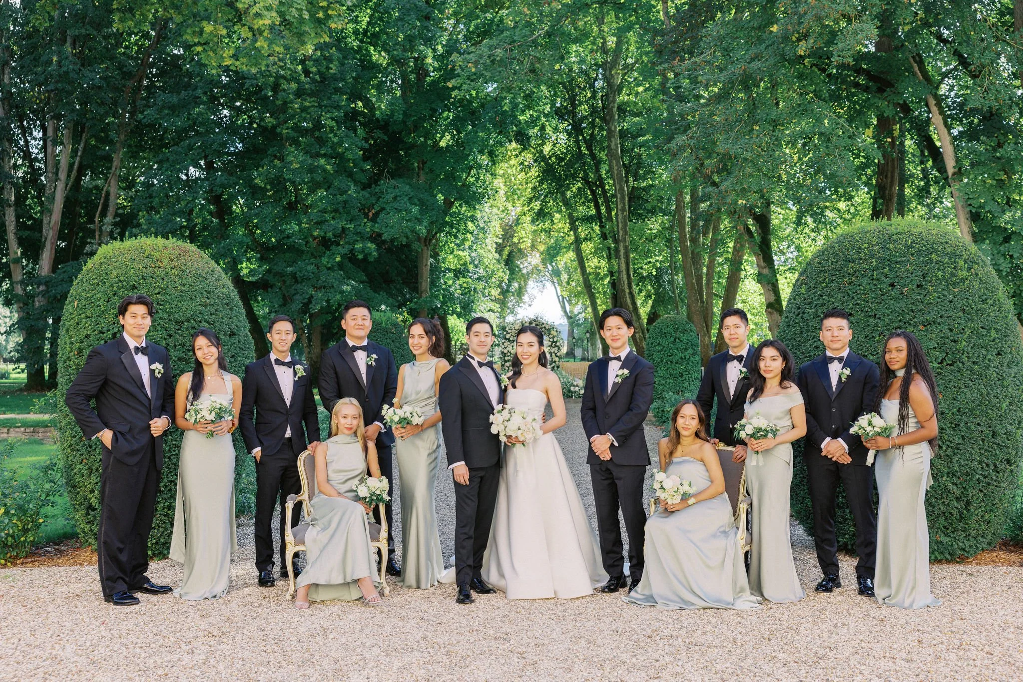 A wedding party with 15 people outdoors in a green park, including a bride and groom at the center, flanked by bridesmaids and groomsmen, all dressed formally and holding flowers.