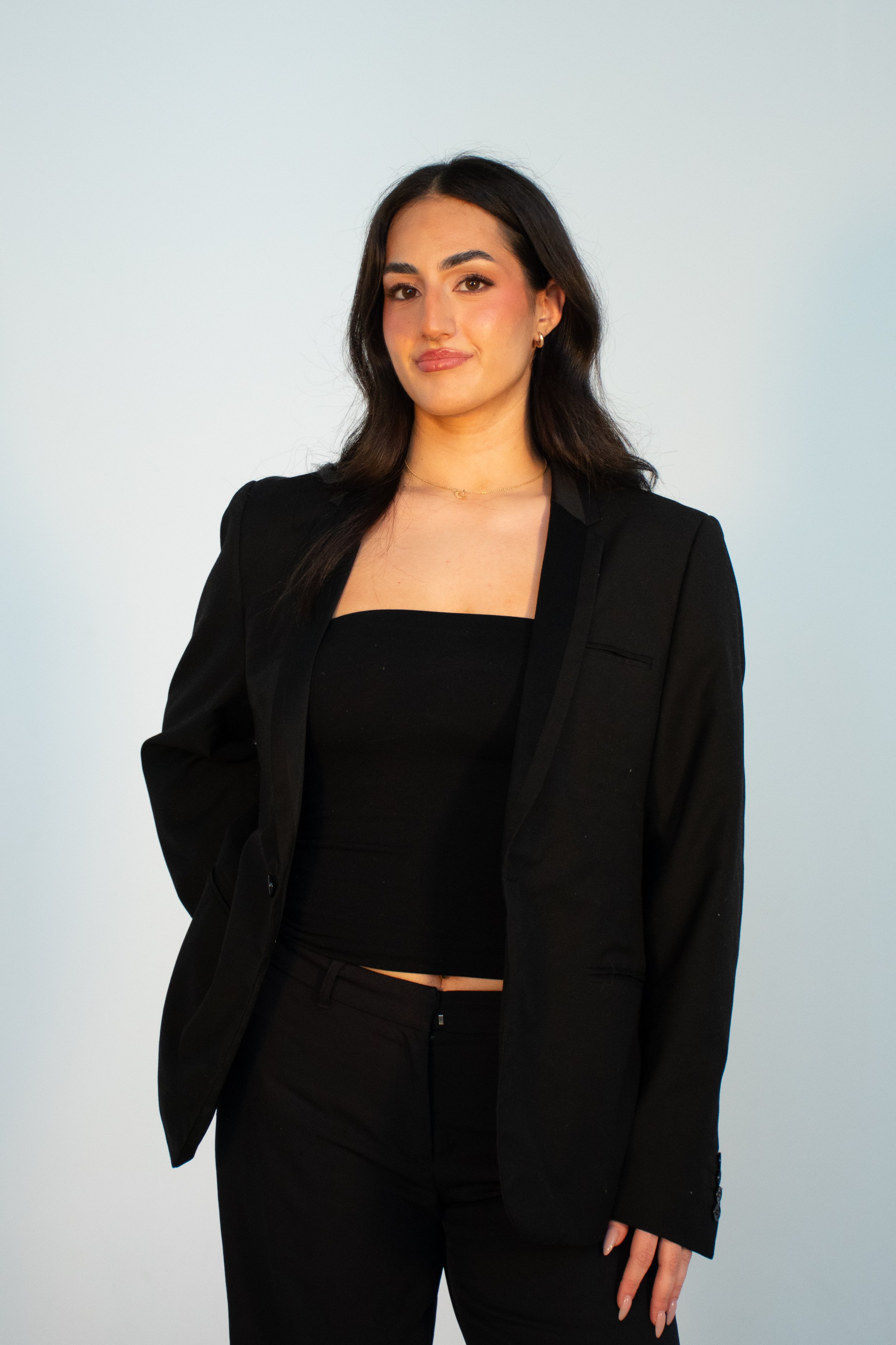 A woman with dark hair wearing a black blazer and black top, standing against a light background.