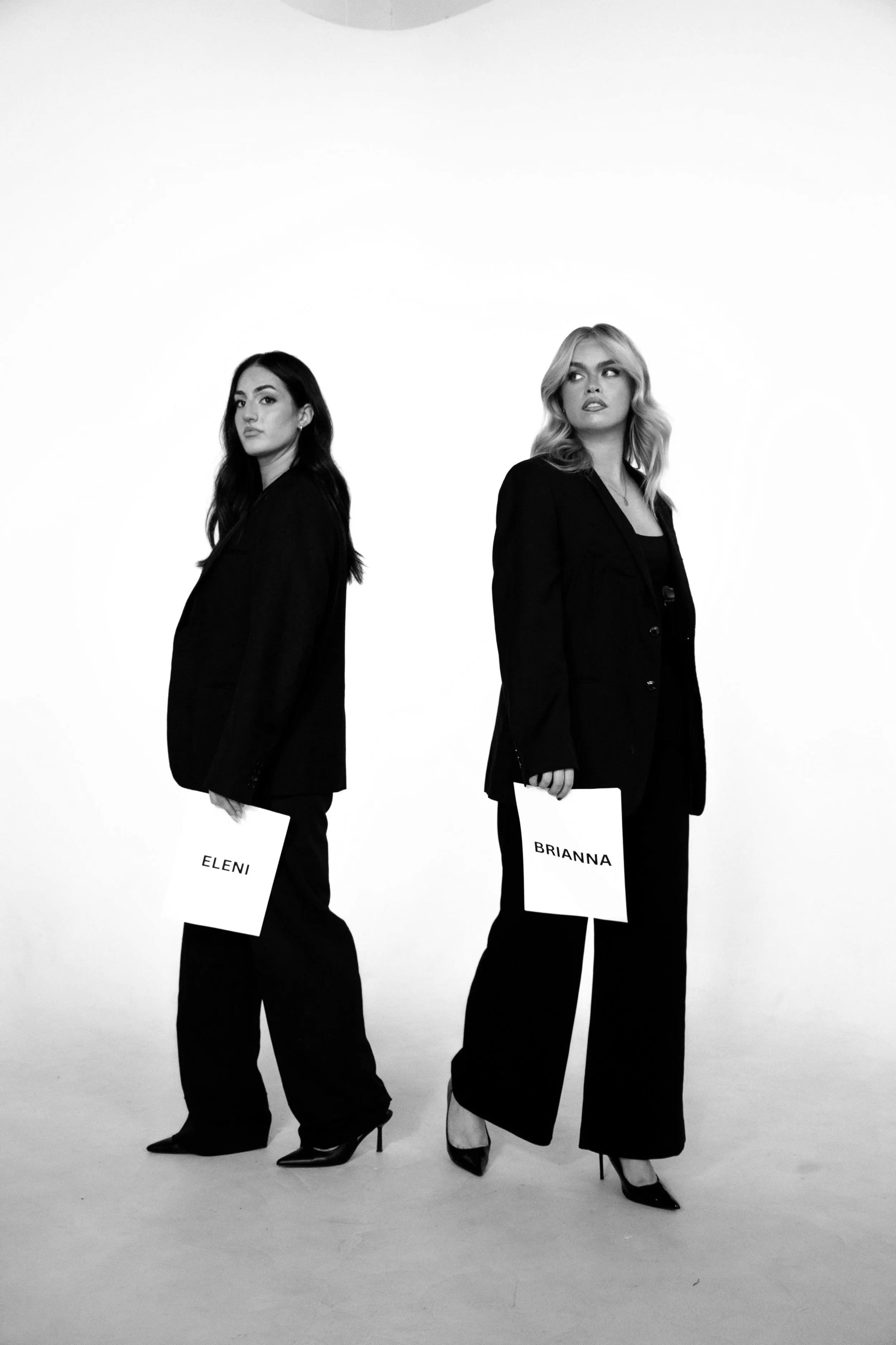 Two women standing in a photo studio, each holding a sign. The woman on the left has dark hair and is holding a sign that says 'ELENI,' the woman on the right has blonde hair and is holding a sign that says 'BRIANNA.' Both are dressed in black suits and wearing high heels, with a plain white background.