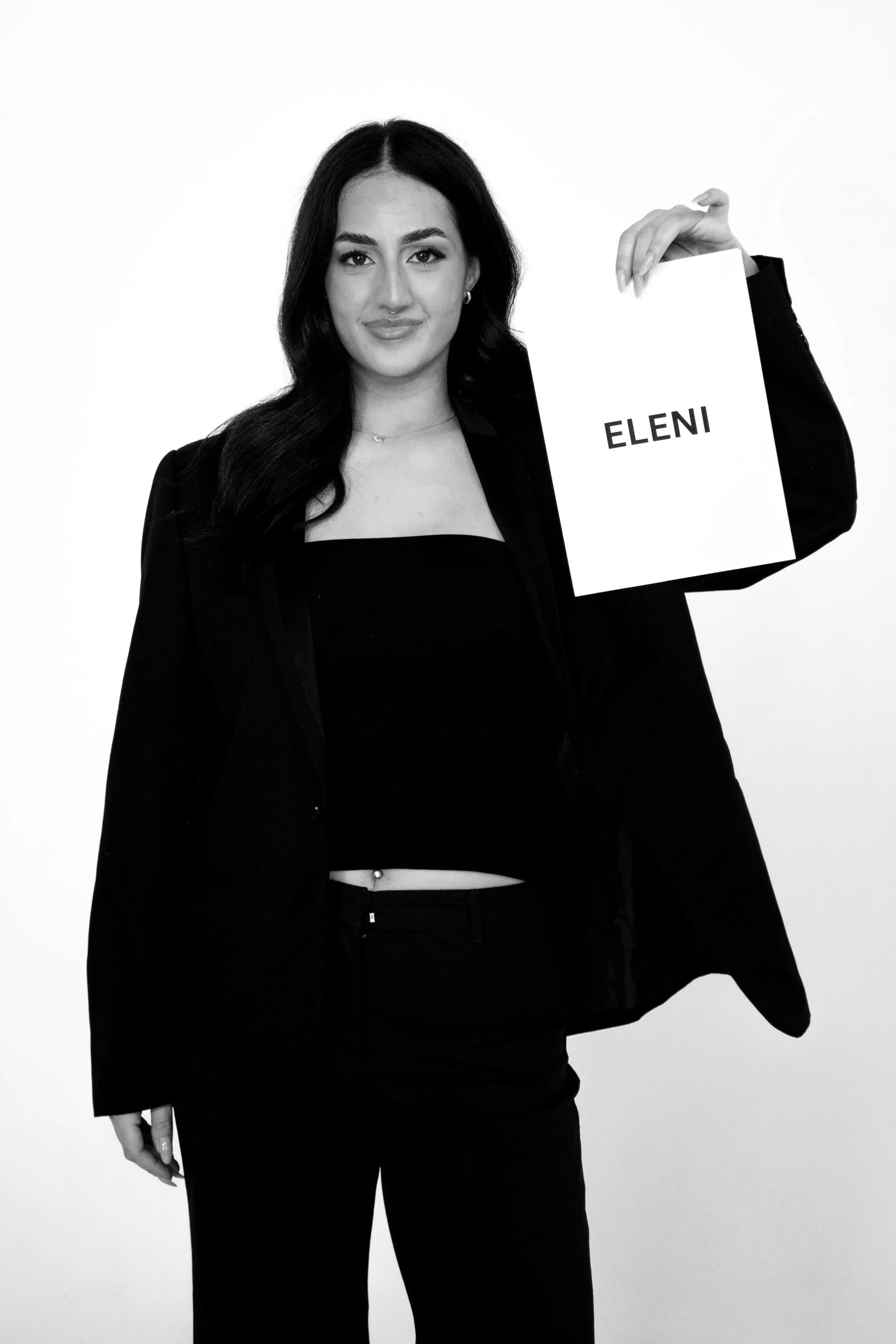 A woman in a black suit holding a sign with the name 'ELENI' written on it, standing against a plain white background.