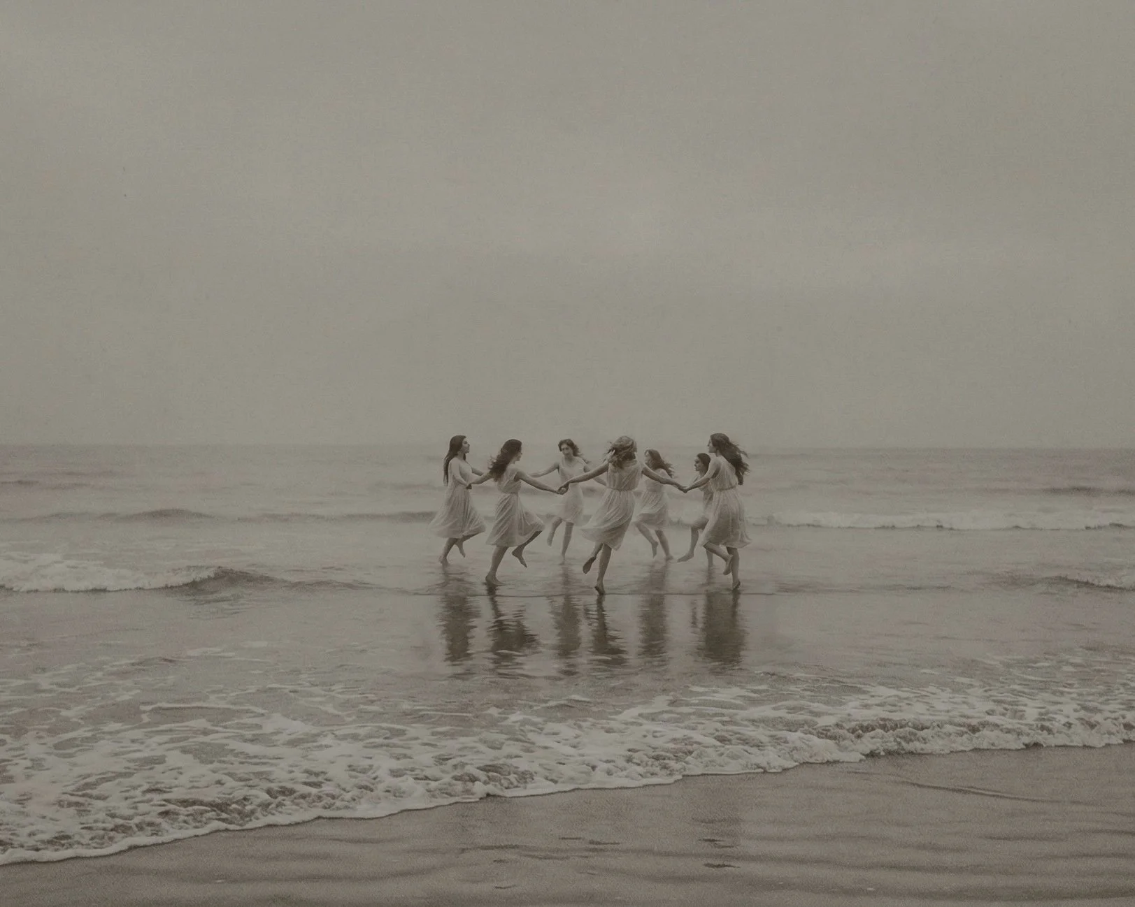 Sieben Frauen in weißen Kleidern tanzen im Wasser am Strand bei Sonnenuntergang.