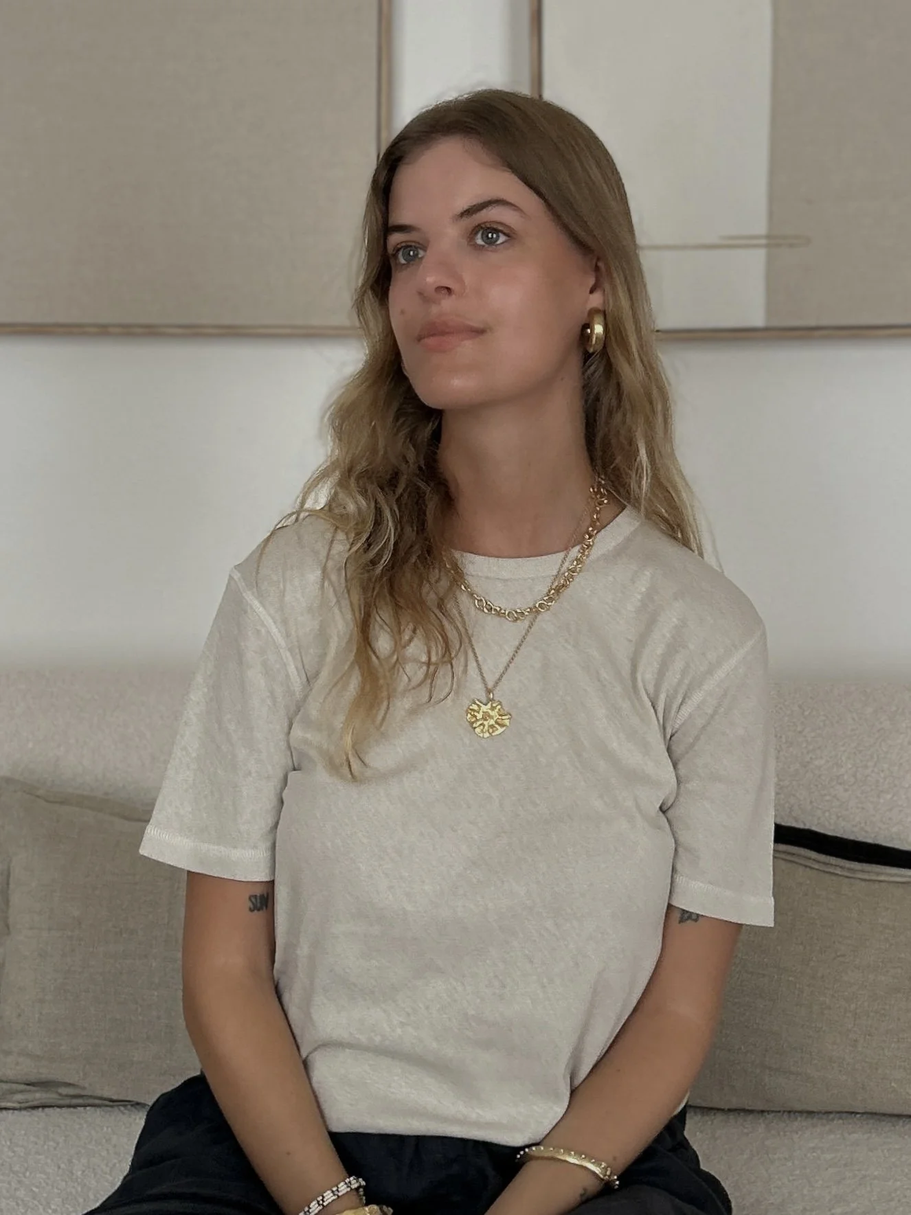 Junge Frau mit blonden, gewelltem Haar, trägt goldene Schmuckstücke und ein helles T-Shirt, sitzt auf einer Couch in einem Raum mit Bildern an der Wand.