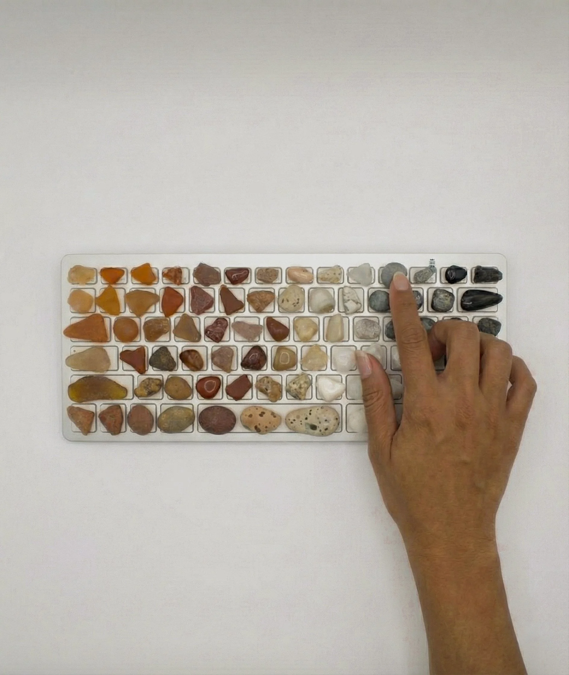 Hand, die mit einem Finger auf eine Tastatur mit verschiedenen bunten Steinen tippt, auf weißem Hintergrund.