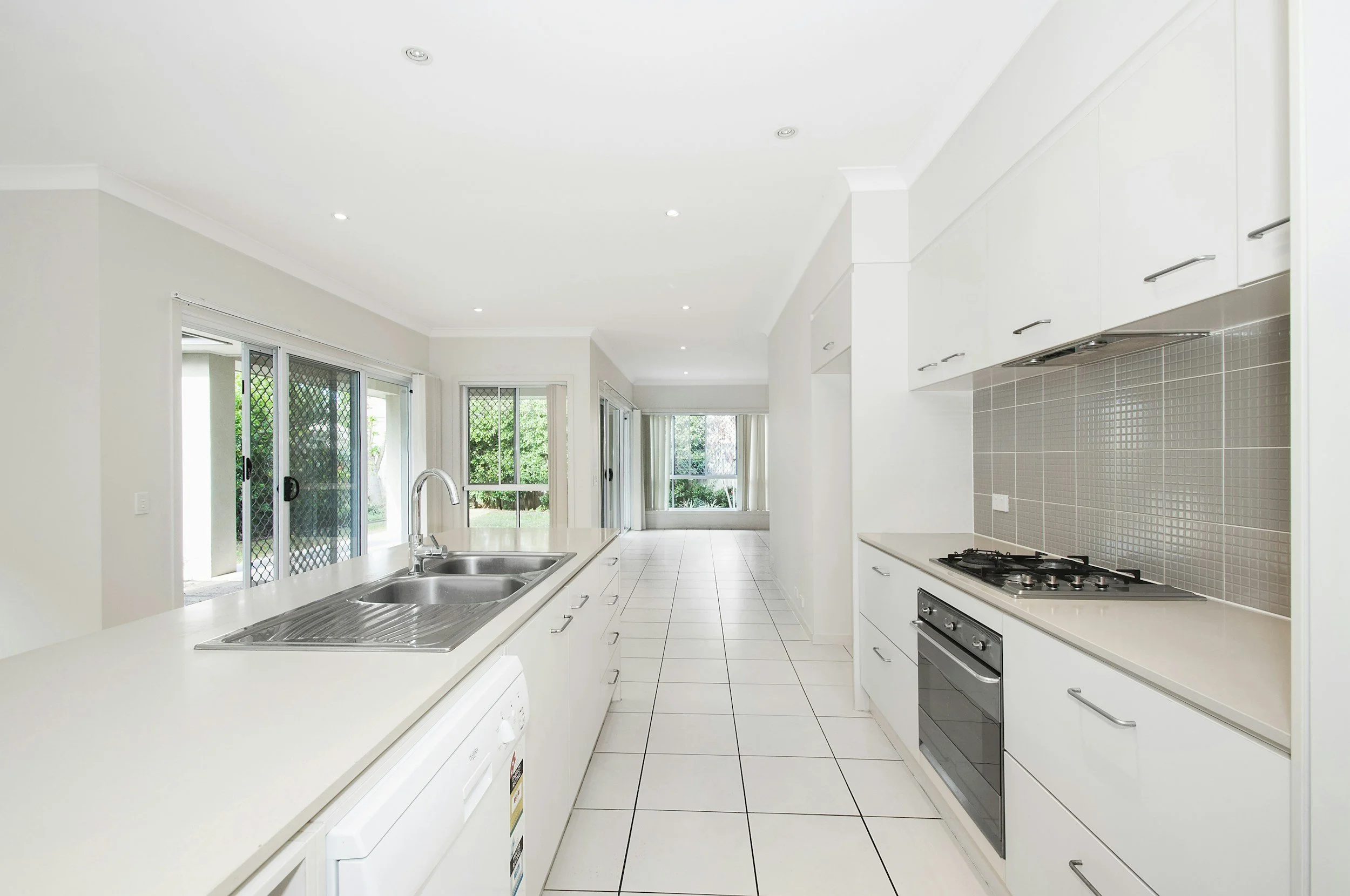 A bright, modern kitchen with white cabinets, a tiled backsplash, and large windows that allow natural light to fill the space. The kitchen features a countertop with a double sink, a gas stove, and tiled floors.