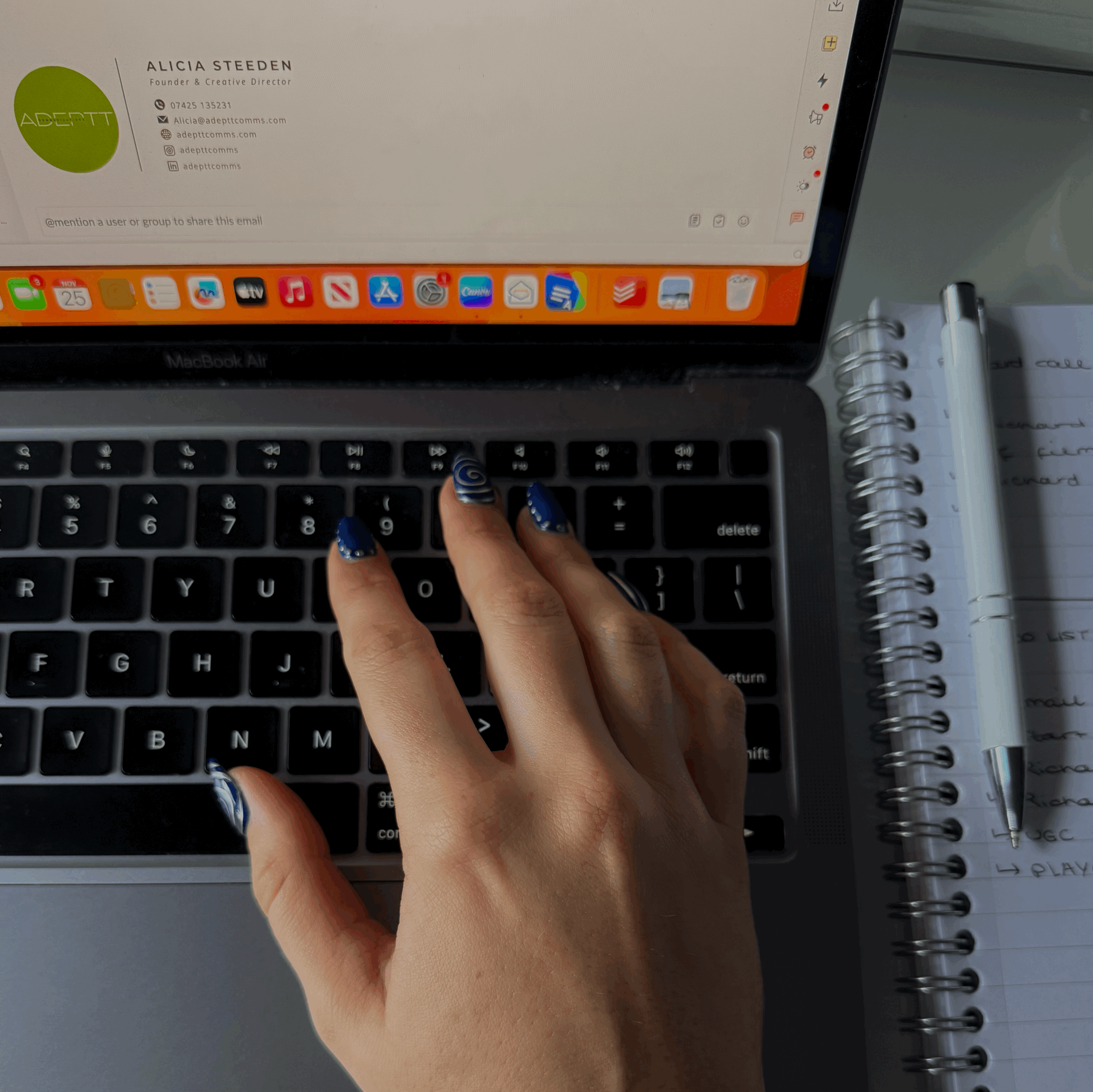 Close-up of a person's hand with decorative blue and white nail art on a MacBook keyboard, with an open notebook and a white pen on the right and an email on the screen.