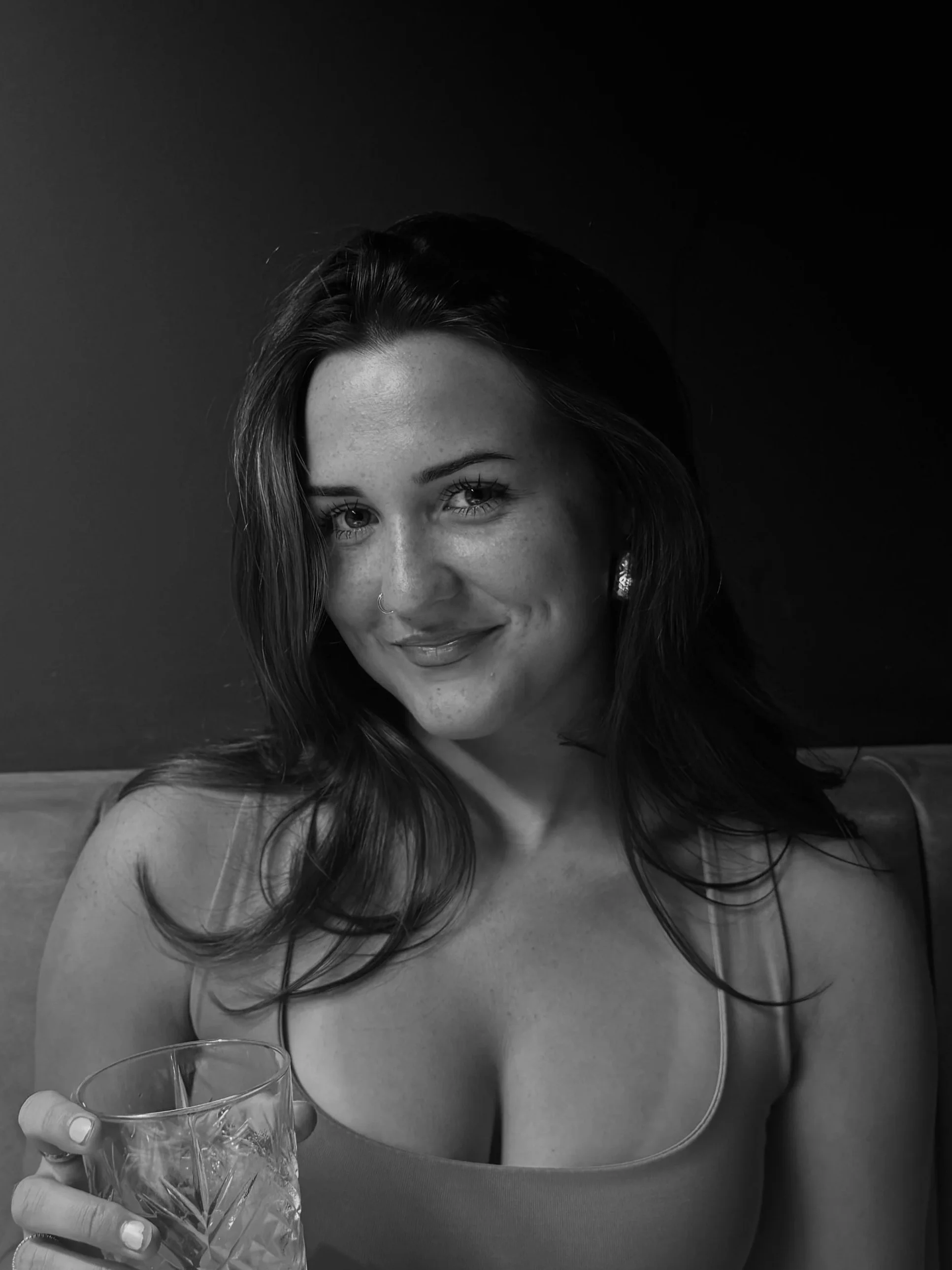 A woman with long dark hair, smiling, holding a glass of drink, wearing a tank top, in a dimly lit setting. Social media manager. Social media management.