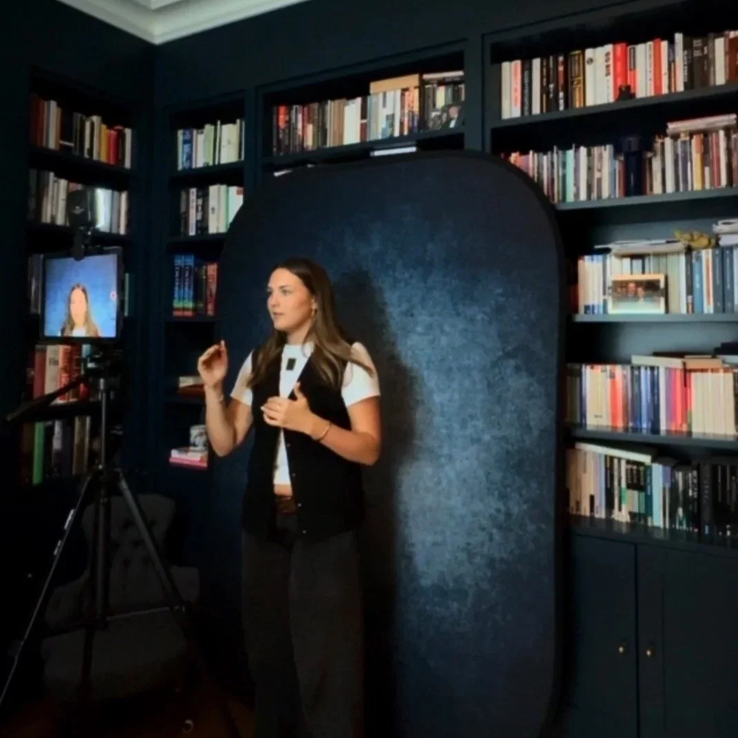 A woman is standing in front of a dark blue bookcase and camera, with a black backdrop behind her, speaking or presenting, in a room filled with books.