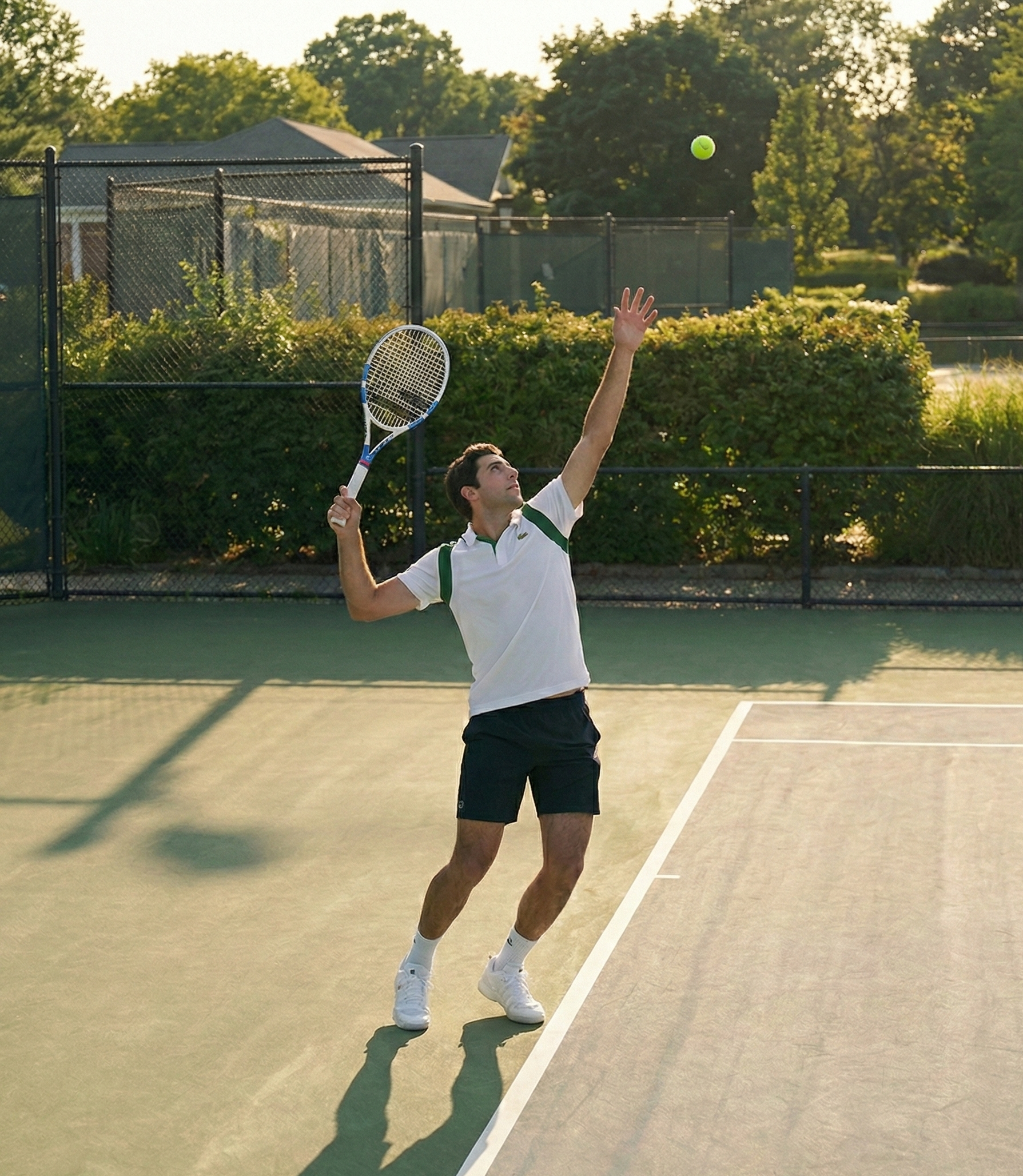 Joueur de tennis en tenue blanche au service sur un court en gazon entouré de haies