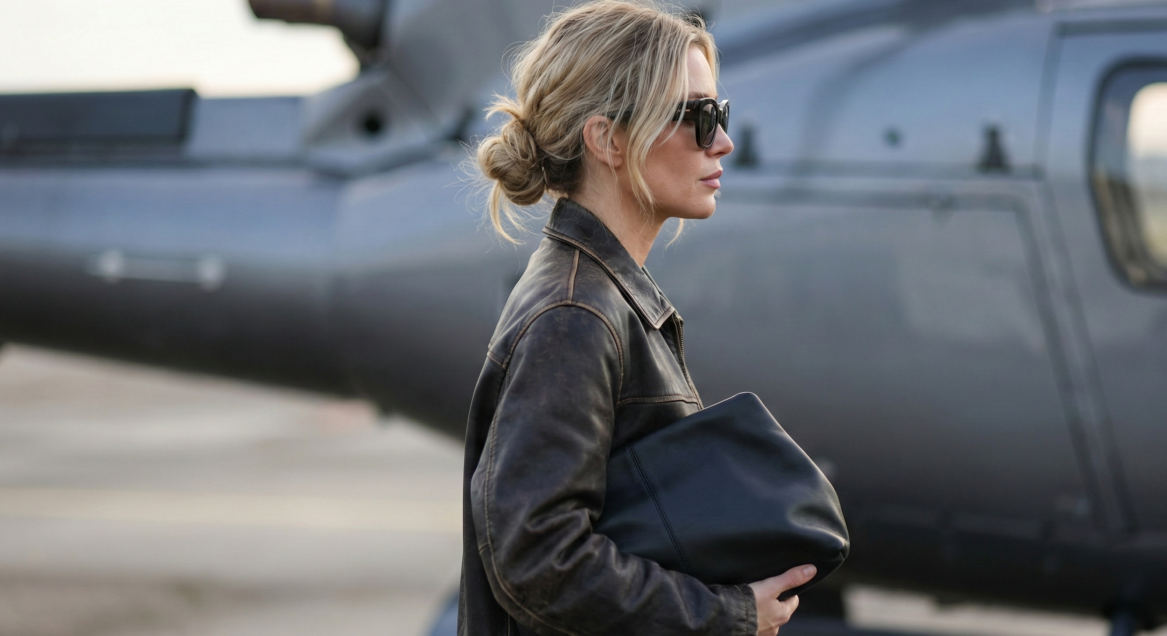 Femme blonde en veste en cuir tenant une pochette noire près d’un hélicoptère