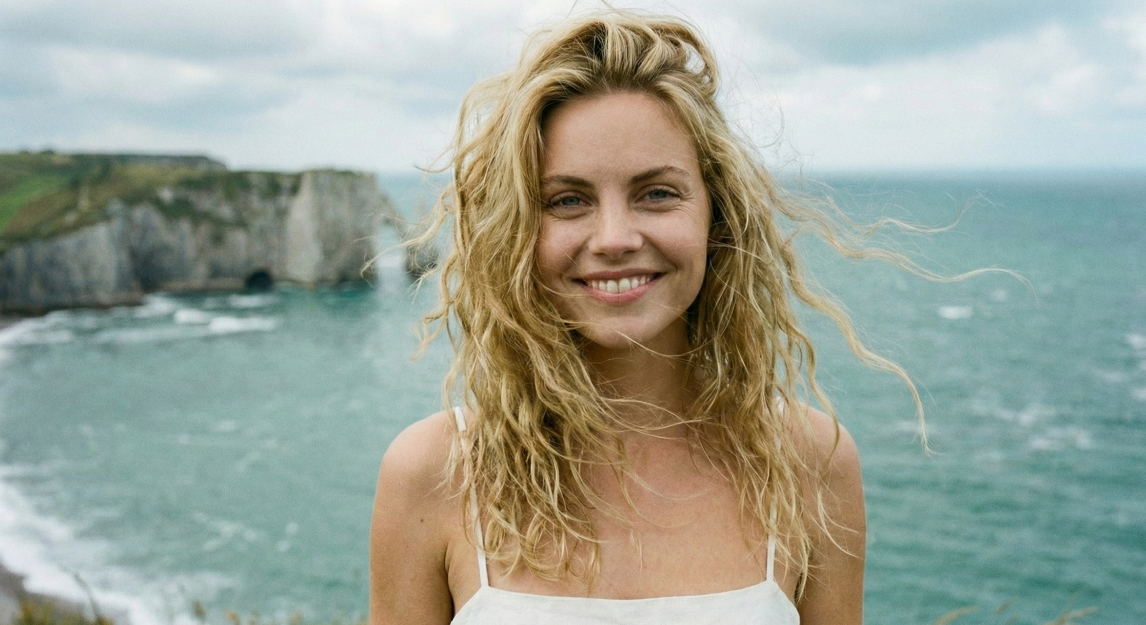 Portrait souriant d’une femme blonde aux cheveux au vent, bord de mer en arrière‑plan