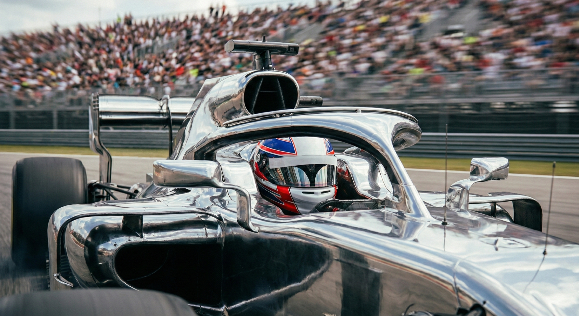 Pilote casqué au volant d’une monoplace argentée, tribunes floues en arrière‑plan