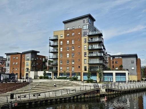 Multi-story apartment buildings along a waterfront with pedestrian walkway and railings, under a cloudy sky.