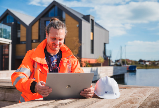 A man in an orange safety jacket looks at a laptop outdoors near a construction site with a white safety helmet on the table.