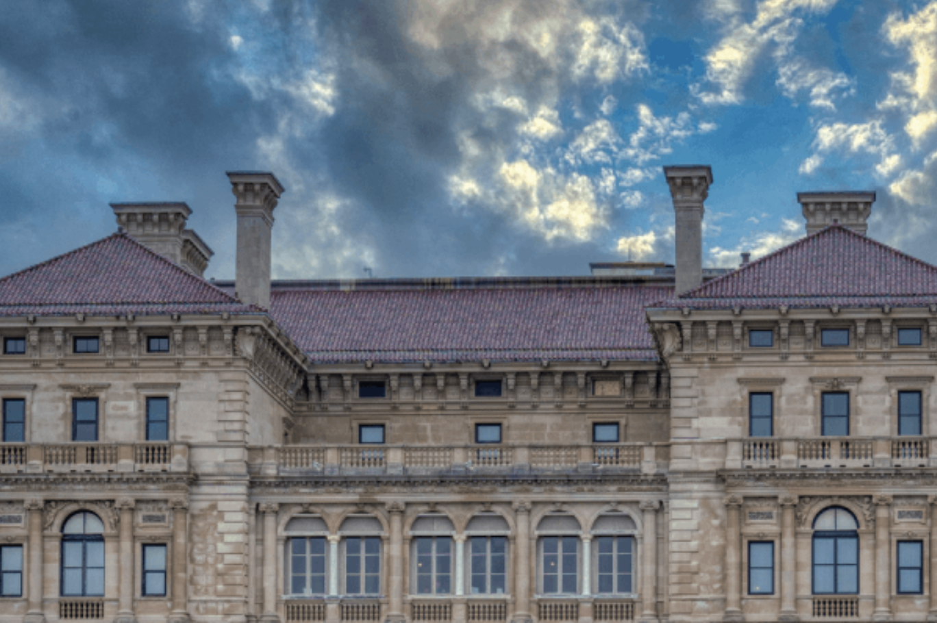Facade of a historic building with beige stone walls, arched windows, and a red tiled roof under a cloudy sky.