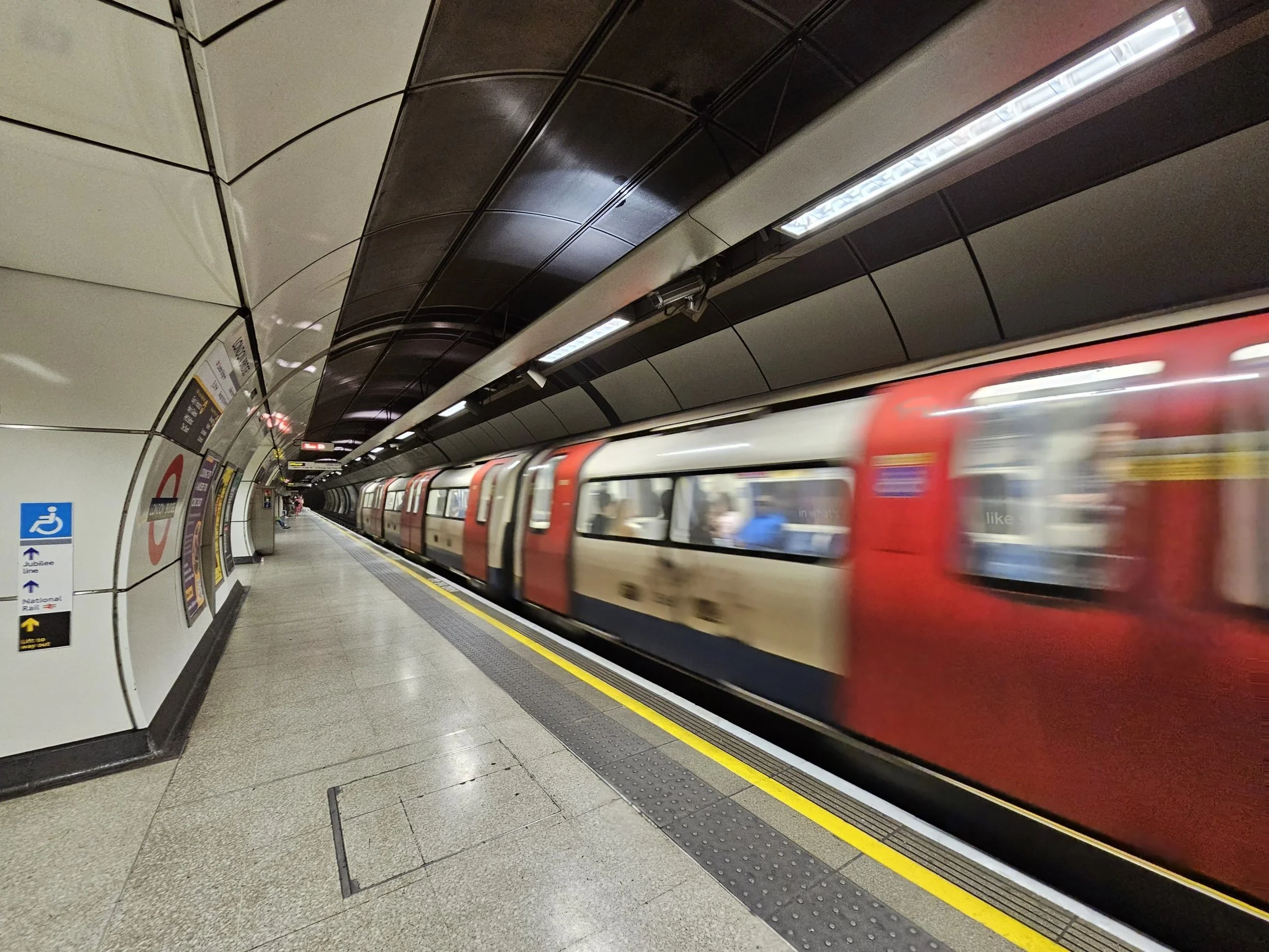London Underground station platform with a passing train, curved tiled wall and signage.