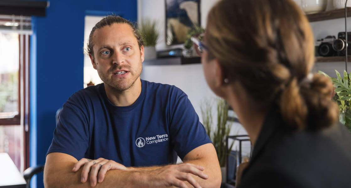 A man wearing a navy blue T-shirt with the logo 'New Terra Compliance' is speaking to a woman with brown hair tied back, sitting indoors with a blue wall and shelves in the background.