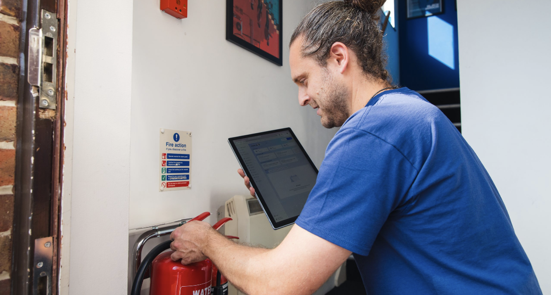 A man in a blue shirt is operating a fire extinguisher while looking at a tablet or laptop.