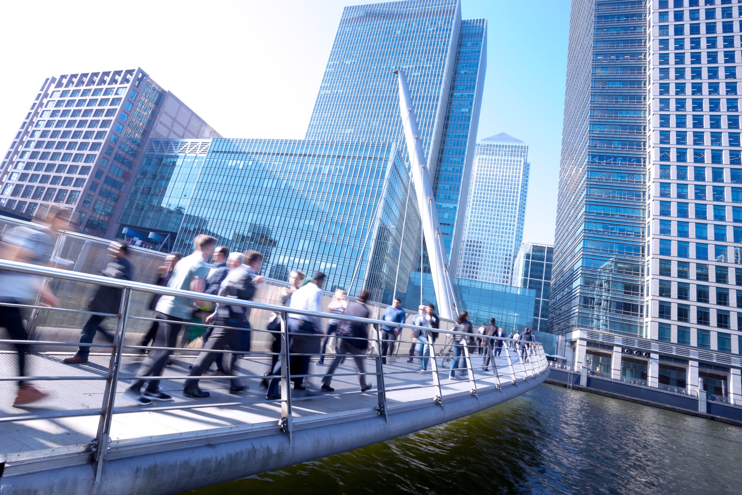 People walking on a modern pedestrian bridge in a city with tall glass skyscrapers reflected in the buildings.