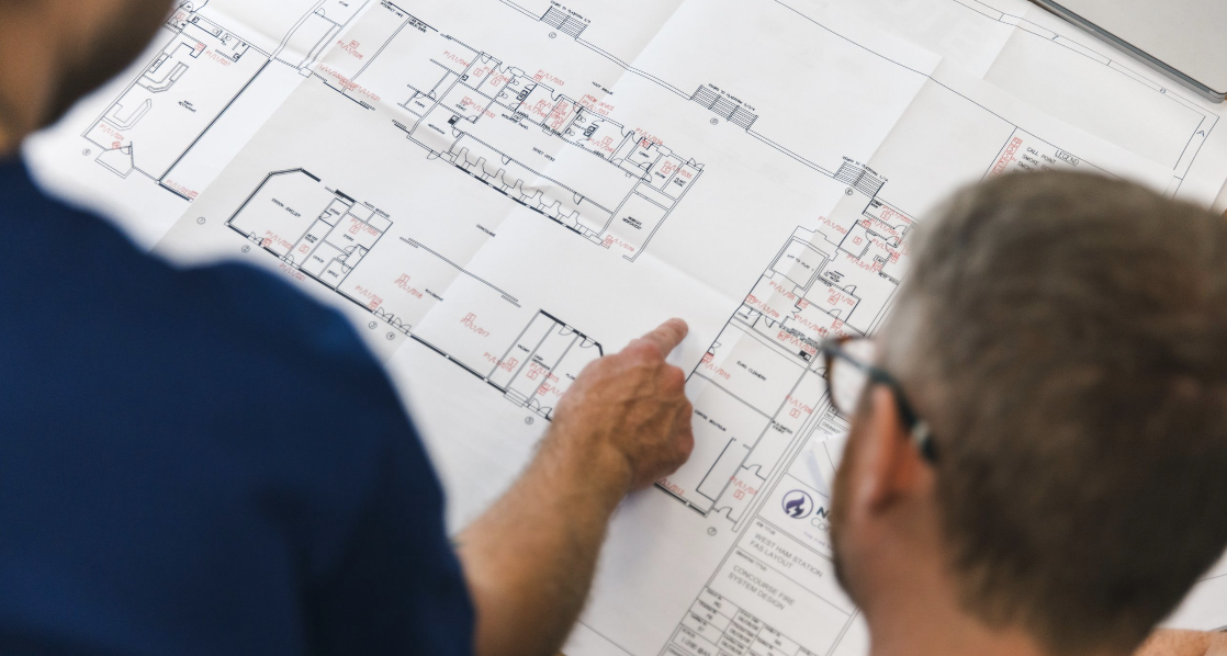 Two individuals reviewing architectural blueprints spread out on a table, with one person pointing at a specific section.