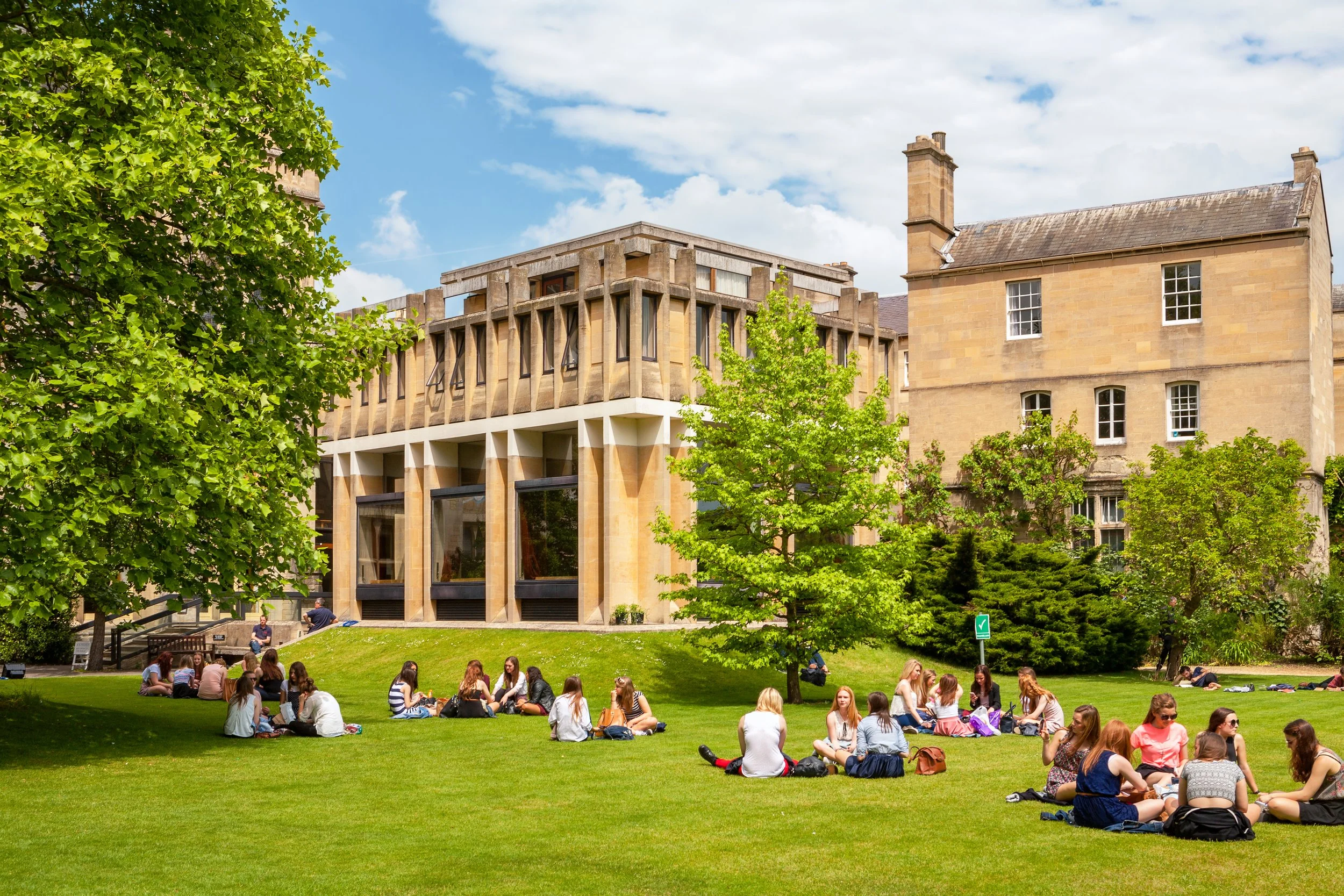 Group of students sitting on grass and talking outside near historic and modern buildings with trees on a sunny day.