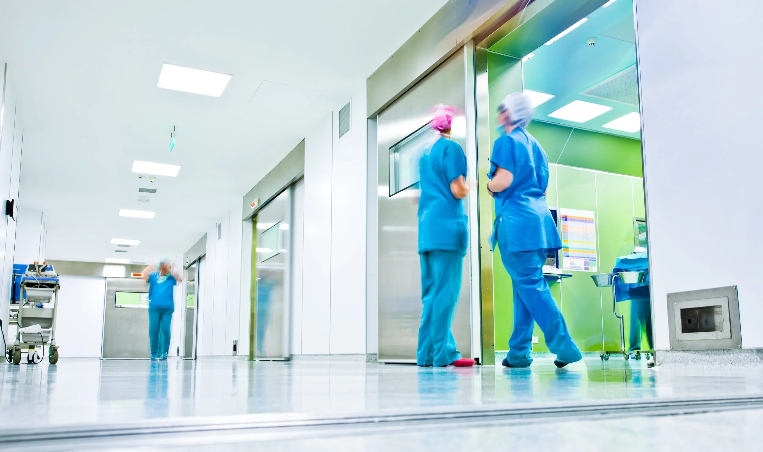 Hospital corridor with three nurses in scrubs and caps, two standing and conversing near an elevator and one walking in the background.