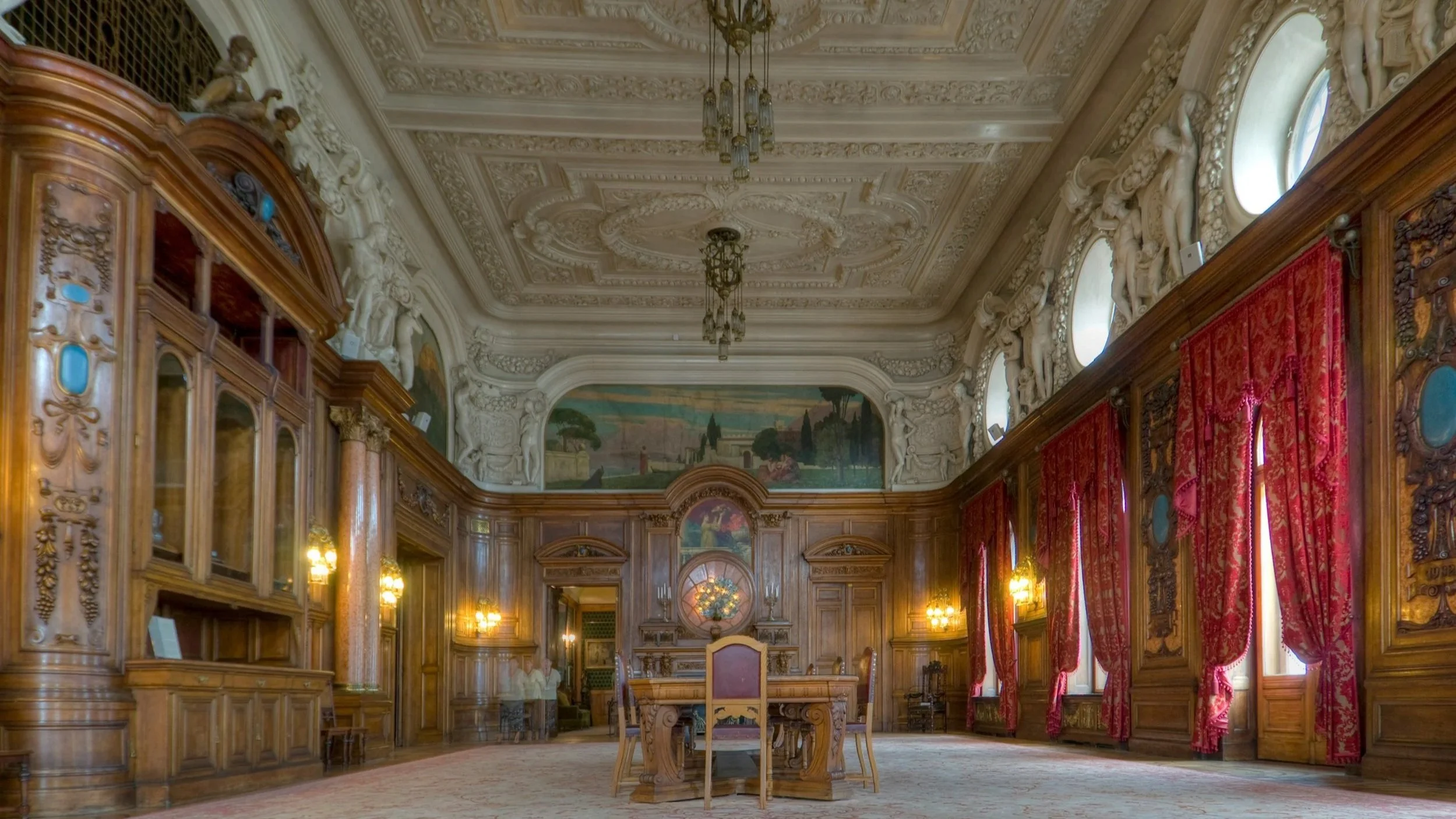 A grand, ornate wooden-paneled room with a high, decorated ceiling, red curtains along the windows, a large painting above the fireplace, and a central wooden table with chairs, chandeliers, and classical sculptures.