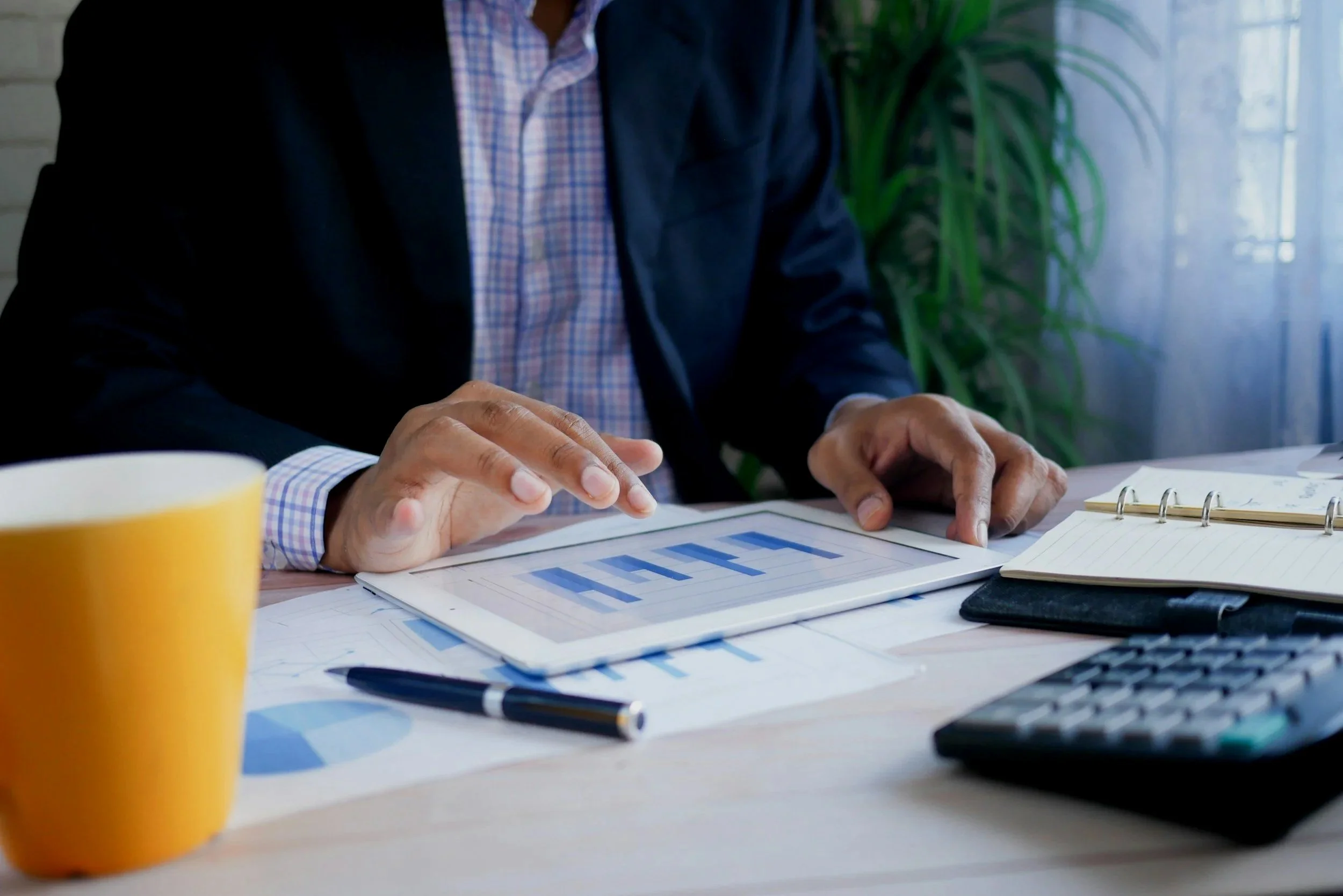 A person in business attire working with a tablet displaying a bar chart, on a desk with a yellow mug, a pen, a notebook, and a calculator, in a well-lit office with a plant in the background.