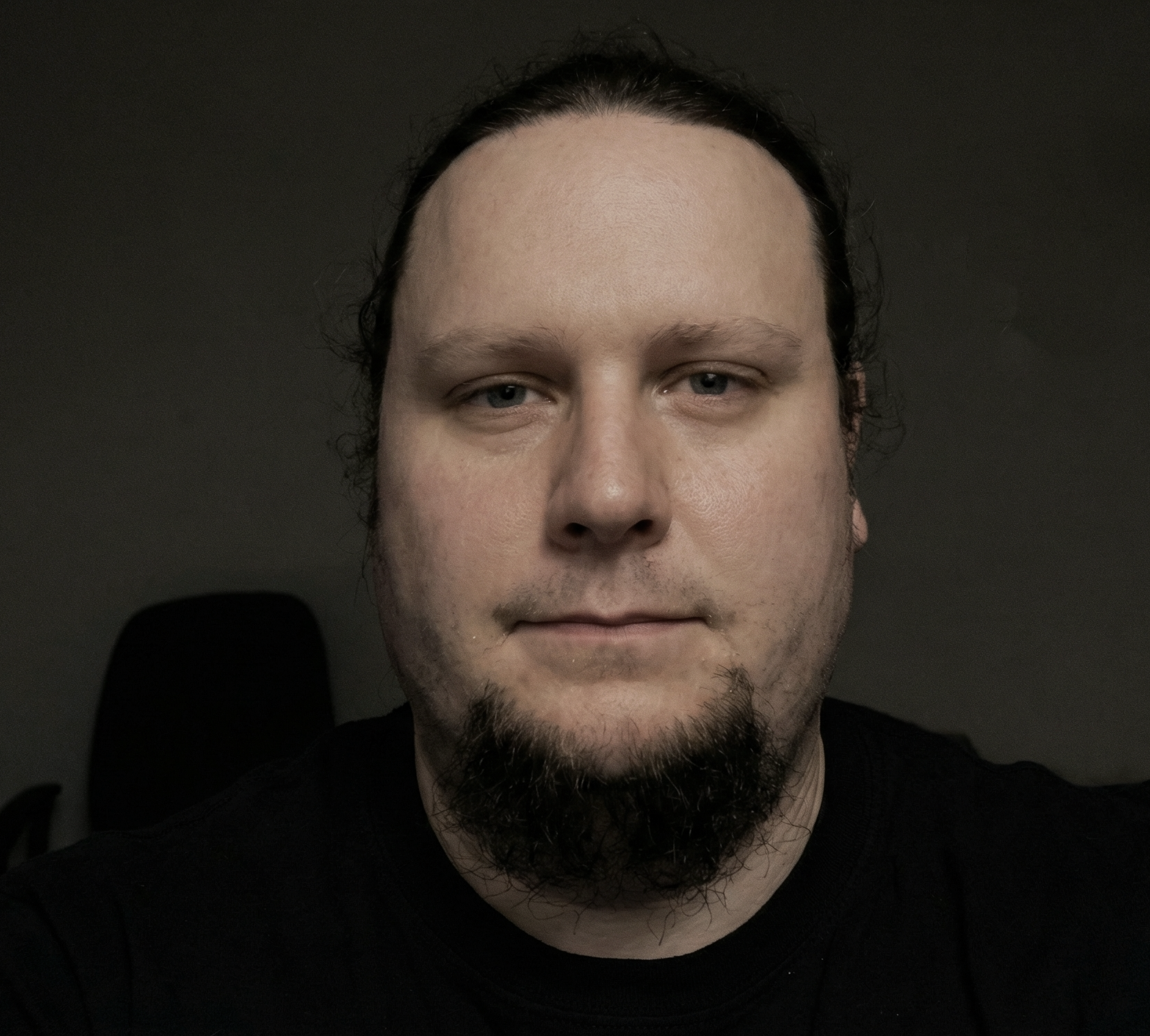 A close-up portrait of a man with light skin, dark hair pulled back, a short dark beard and mustache, wearing a black shirt, in a dimly lit room with a dark background.